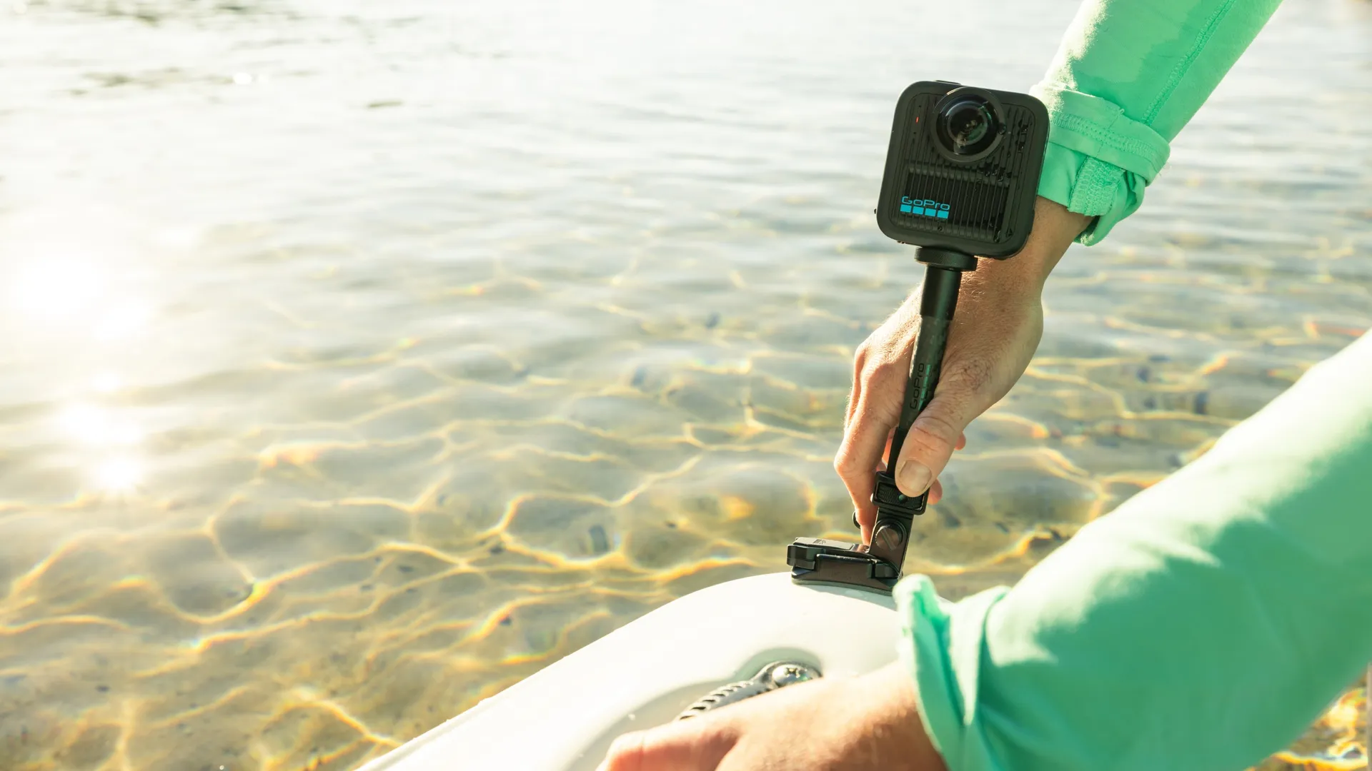 Une personne tient une caméra GoPro près de l'eau. L'eau reflète la lumière du soleil. La personne porte une chemise verte. Un objet blanc est au premier plan.