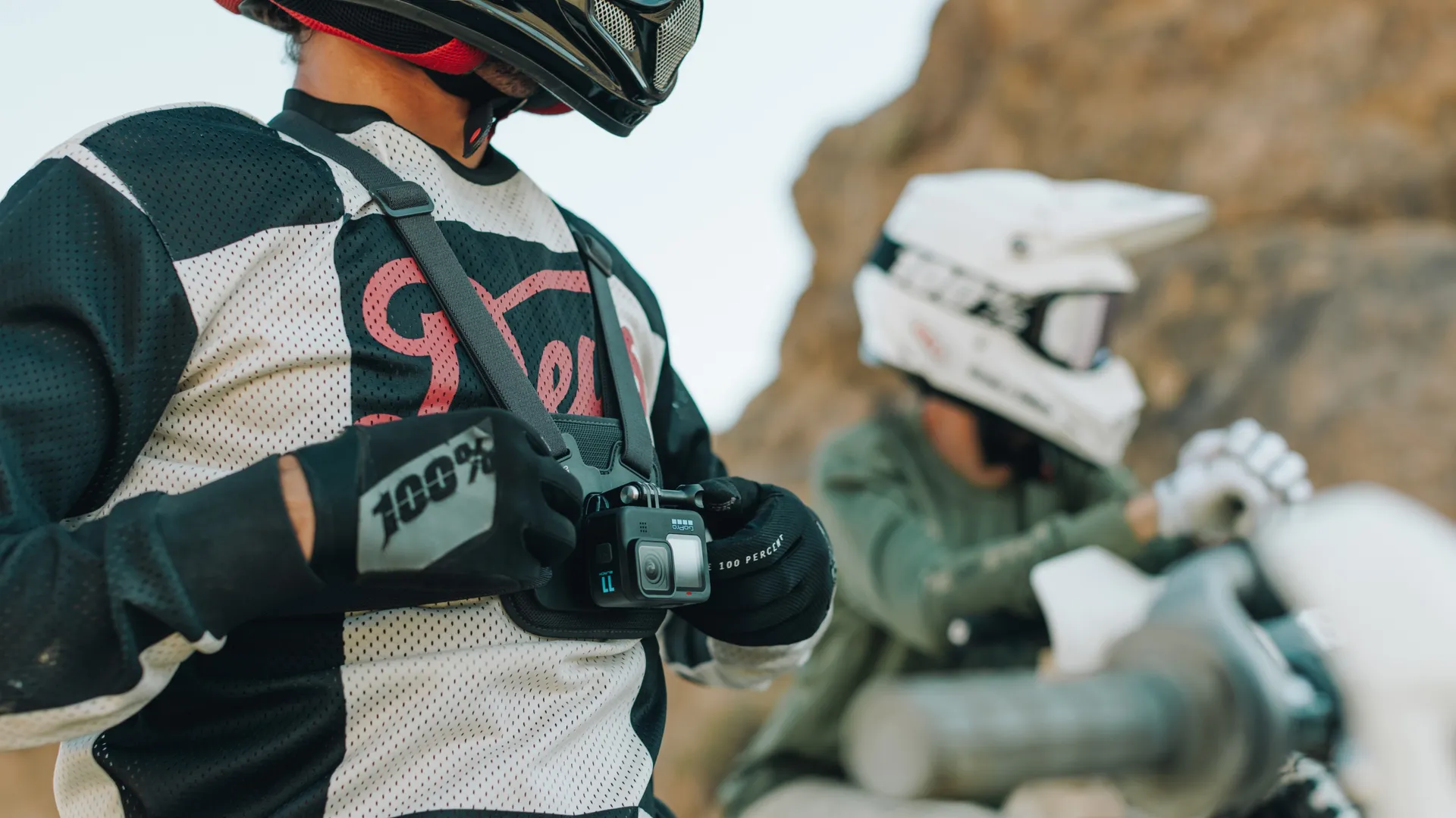 Eine Person mit Helm und Ausrüstung hält eine GoPro-Kamera. Eine weitere Person mit Helm ist im Hintergrund verschwommen, in der Nähe eines Motorrads.