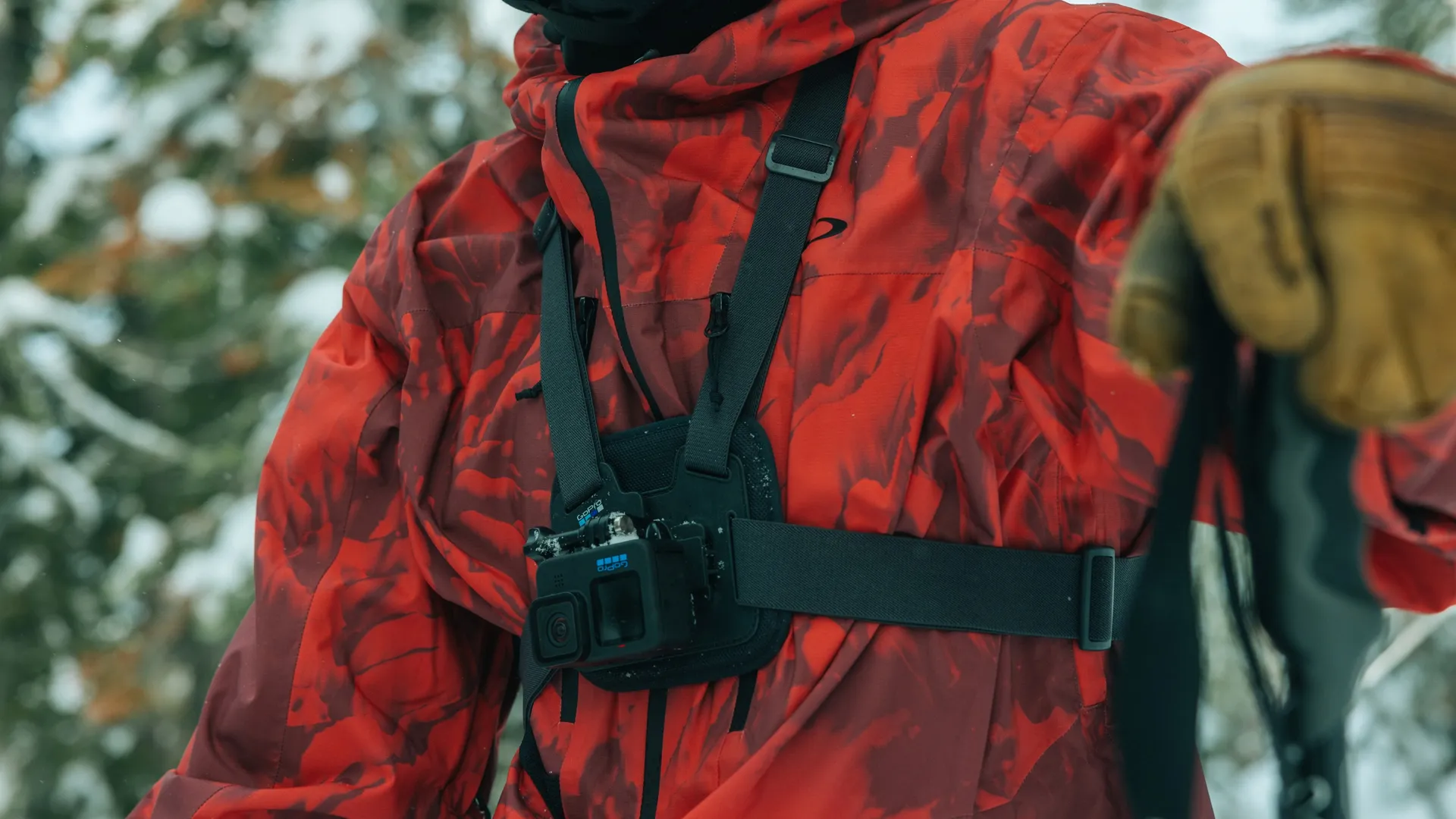 Person trägt eine rot gemusterte Jacke mit einer Brust-GoPro. Skistöcke sind sichtbar, der Hintergrund ist ein verschneiter Wald.