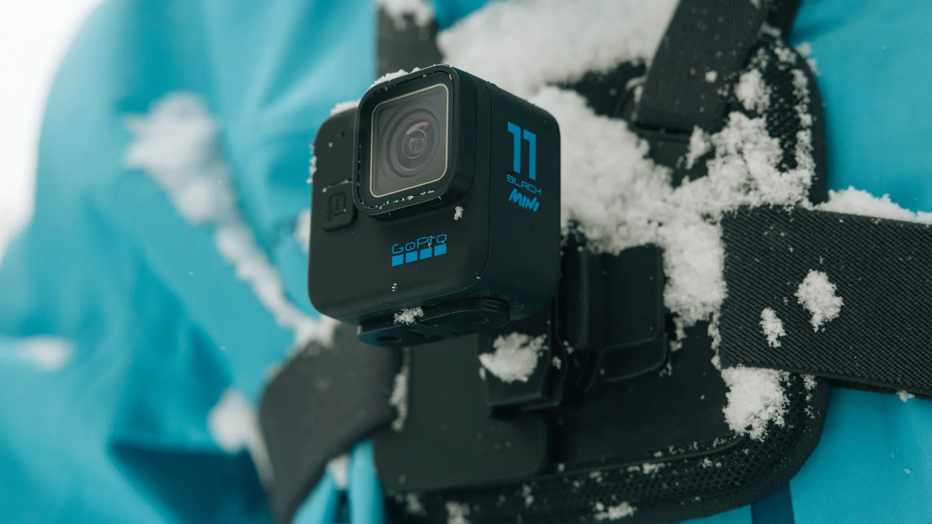 Eine schwarze GoPro 11 Mini Kamera ist an einem Brustgurt befestigt, mit Schnee bedeckt, vor blauem Jackenhintergrund.