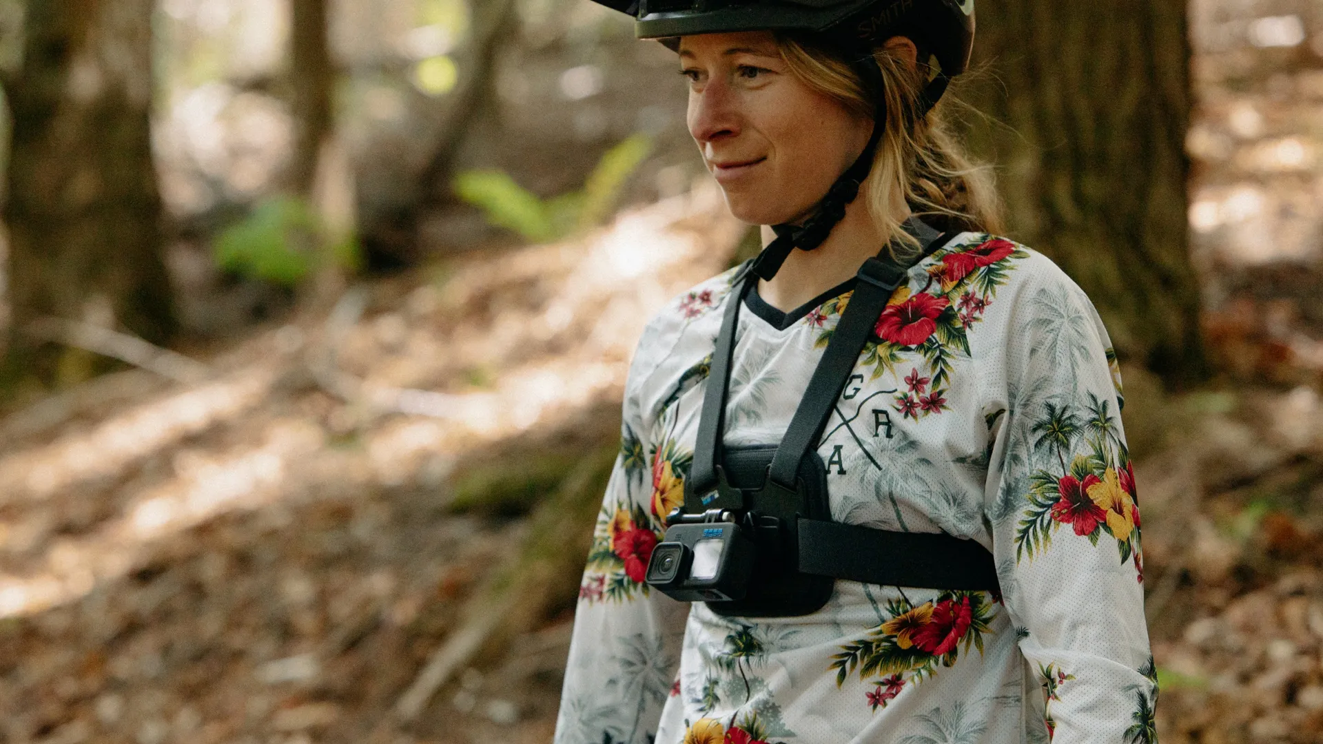 Frau mit Helm und Blumenhemd mit Brustkamera in einer Waldumgebung.