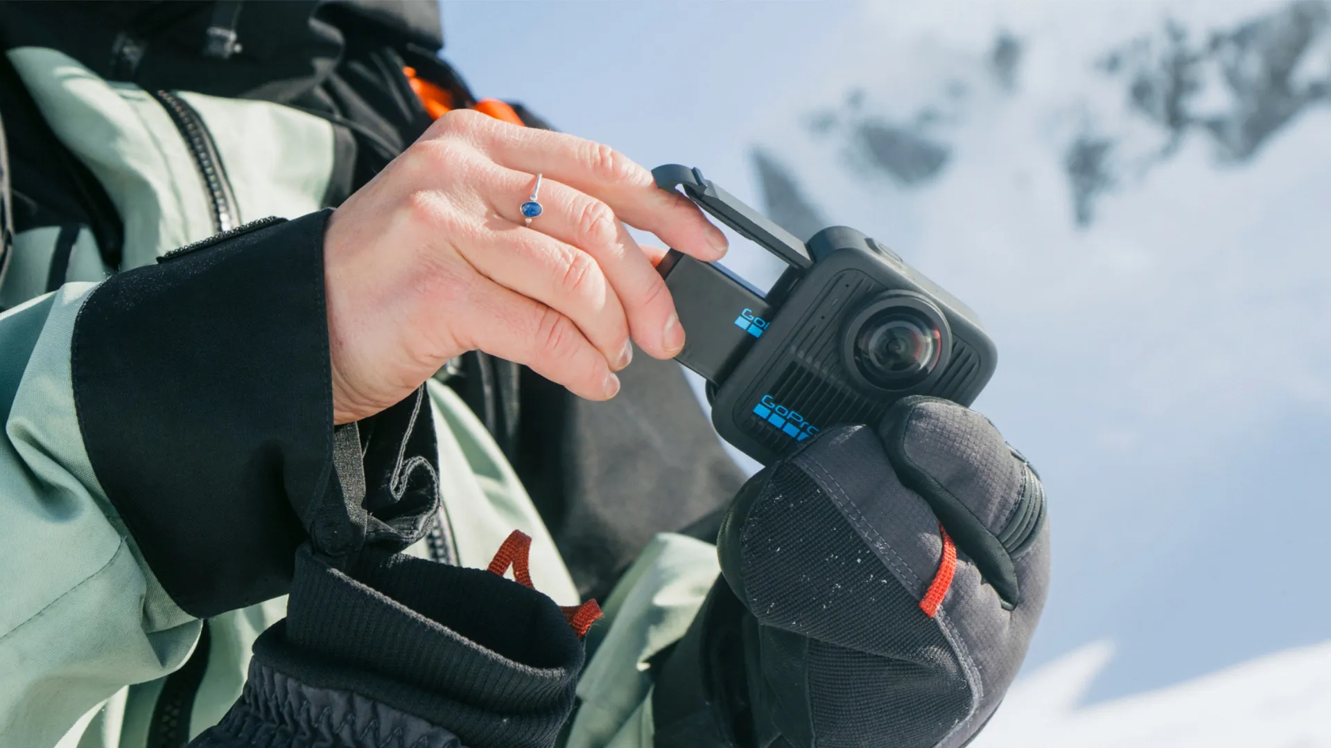 Eine Person hält eine GoPro-Kamera mit behandschuhten Händen. Im Hintergrund ist ein schneebedeckter Berg zu sehen. Die Kamera ist schwarz und blau.
