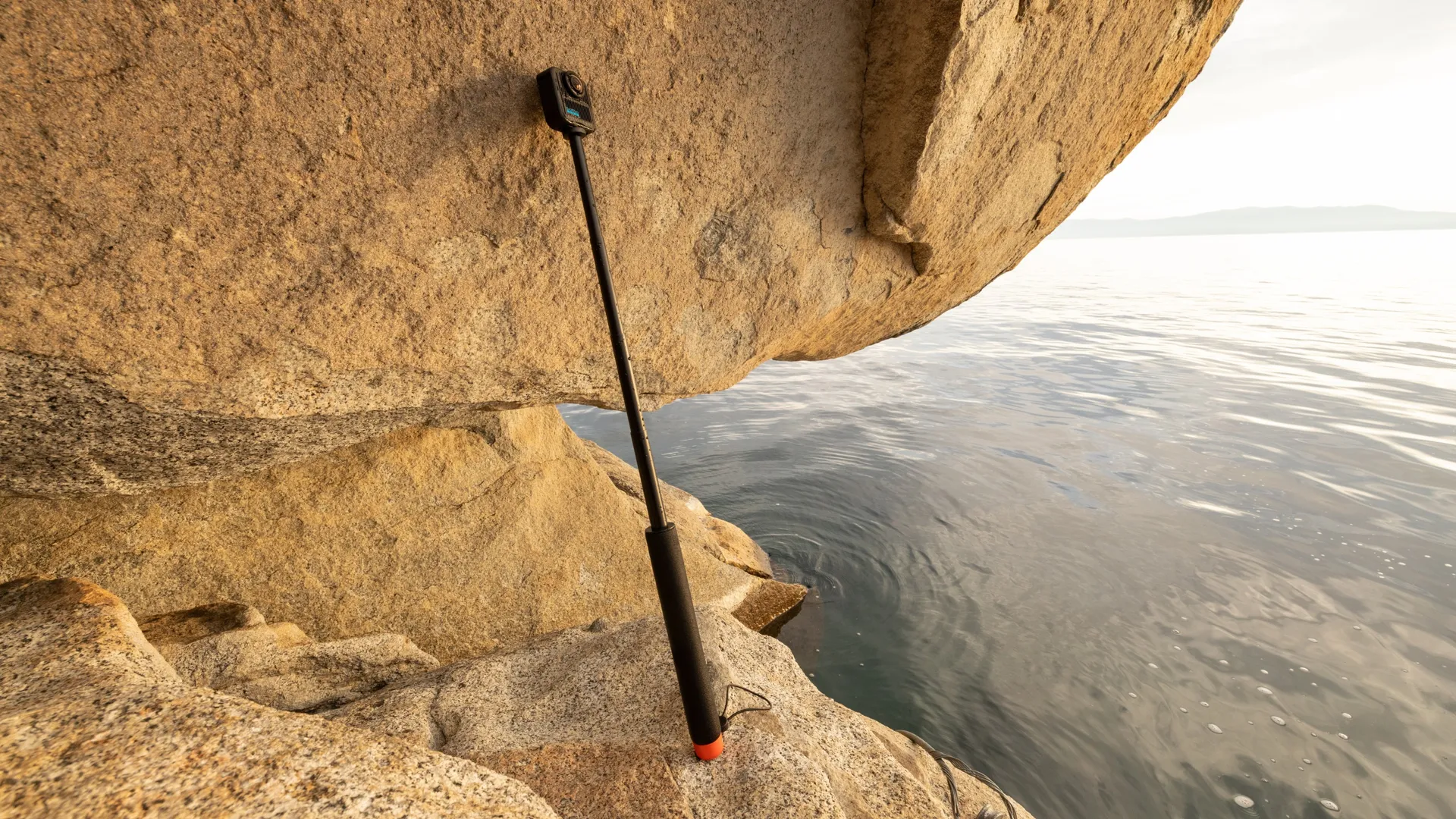 Eine Kamera auf einem Selfie-Stick ist an einer Felsformation befestigt. Ruhiges Wasser und ein entferntes Ufer sind im Hintergrund.
