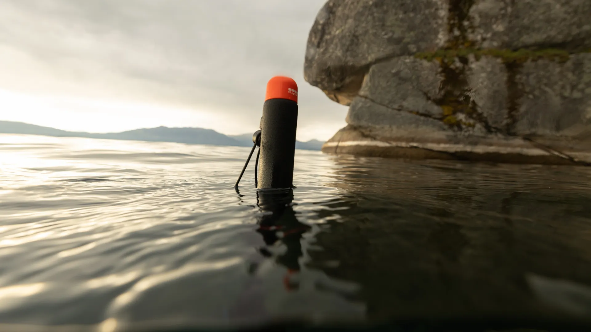 Ein schwarz-oranges Gerät schwimmt im Wasser. Ein großer Felsen und Berge sind im Hintergrund. Der Himmel ist bewölkt.