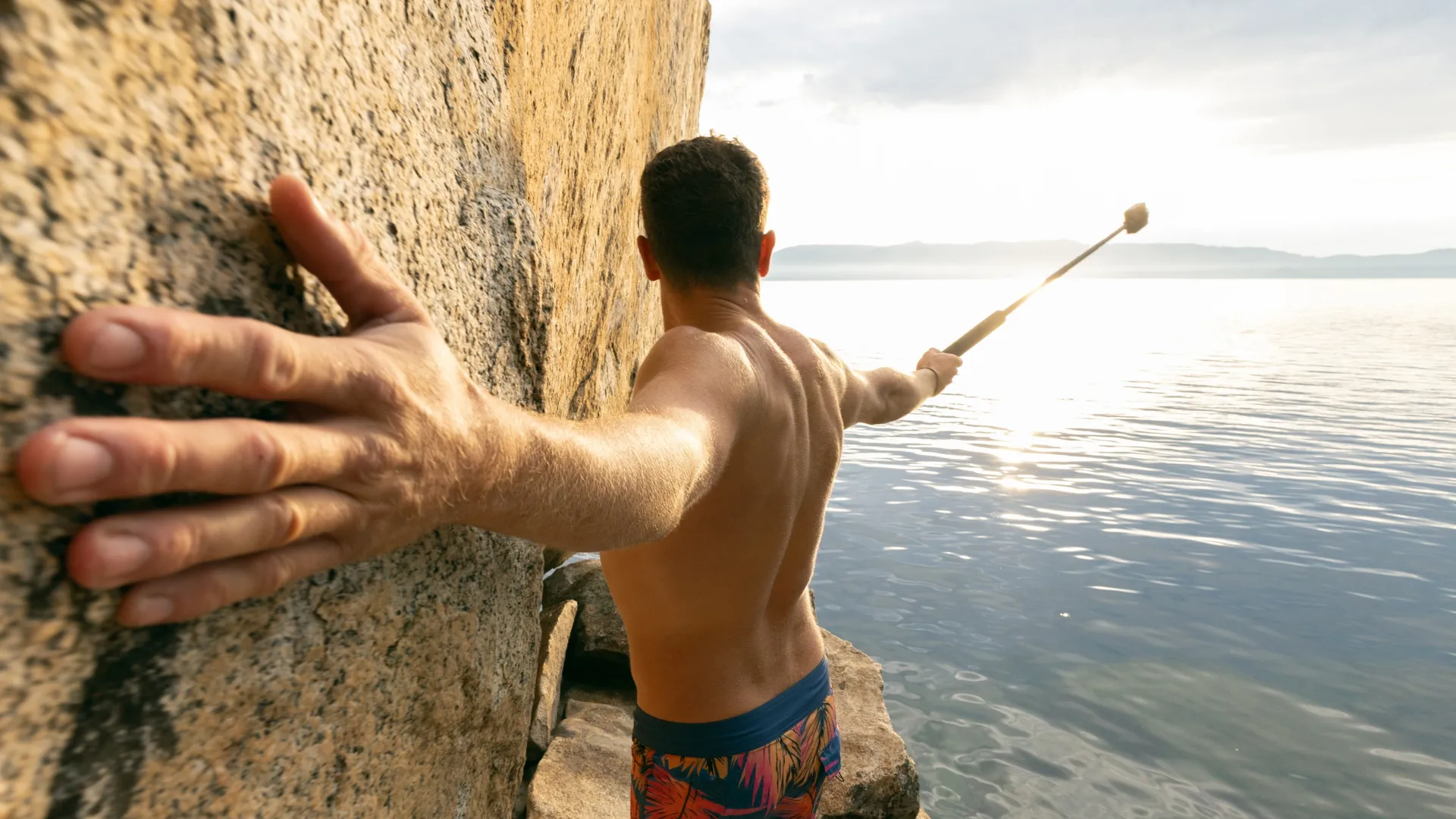 Mann mit ausgestreckten Armen in der Nähe von Felsen und Wasser, hält einen Stock. Blaue Badehose mit floralem Muster.
