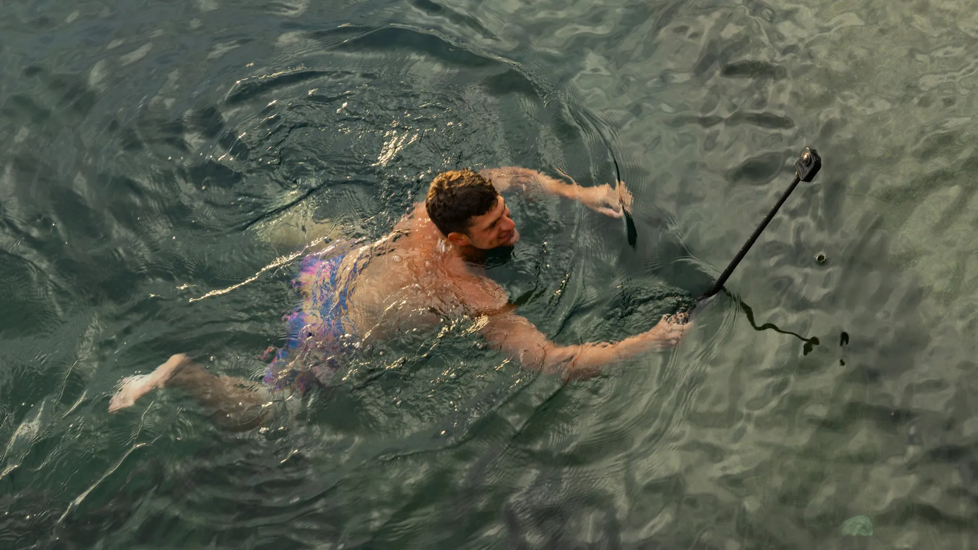 Ein Mann schwimmt in dunklem Wasser und hält einen Selfie-Stick. Er trägt Badehosen und ist teilweise untergetaucht. Das Wasser hat Kräuselungen.
