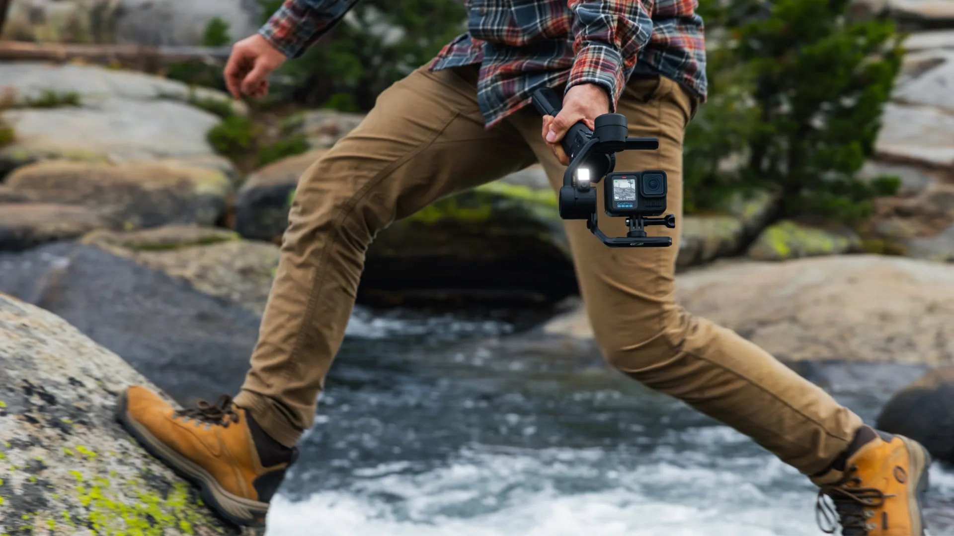 Eine Person springt über einen Bach und hält eine Kamera auf einem Gimbal. Sie trägt ein kariertes Hemd, khakifarbene Hosen und braune Stiefel.