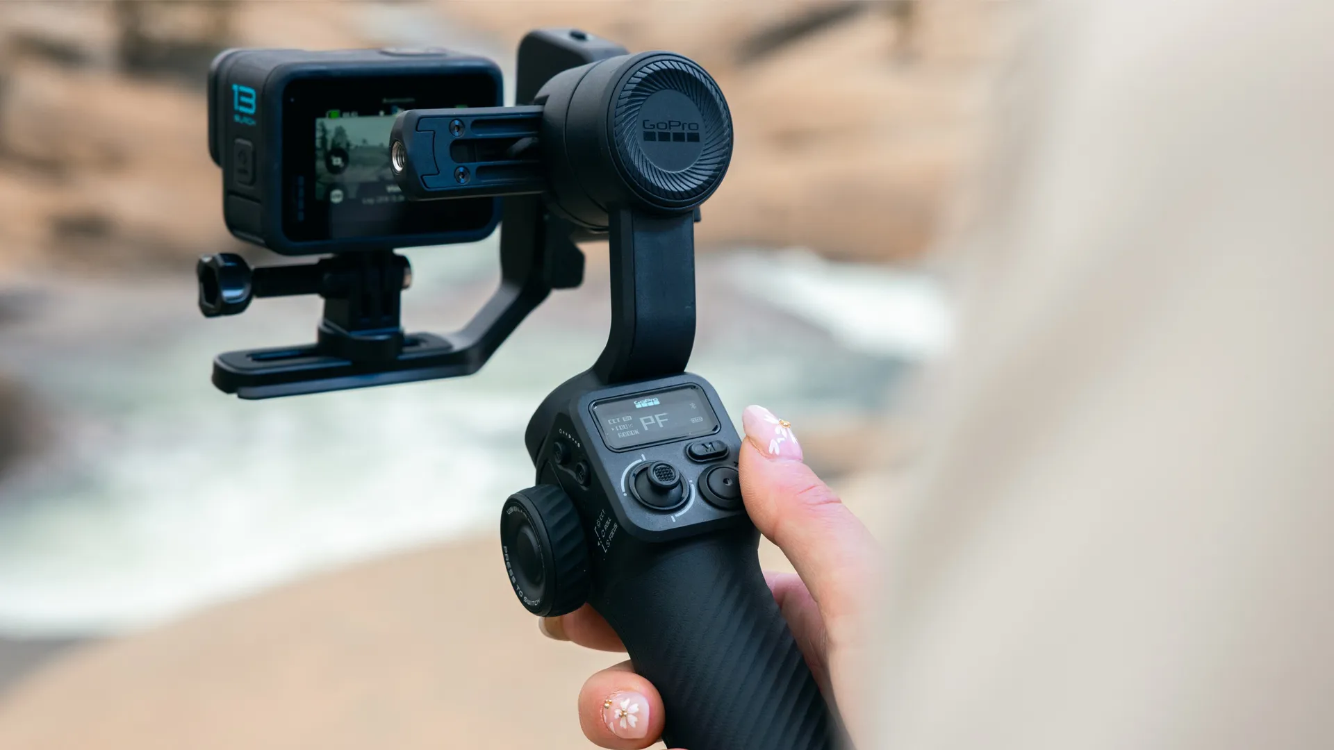 Eine Person hält eine schwarze GoPro-Kamera auf einem Gimbal. Der Kamerabildschirm zeigt eine Szene im Freien. Der Hintergrund ist unscharf.