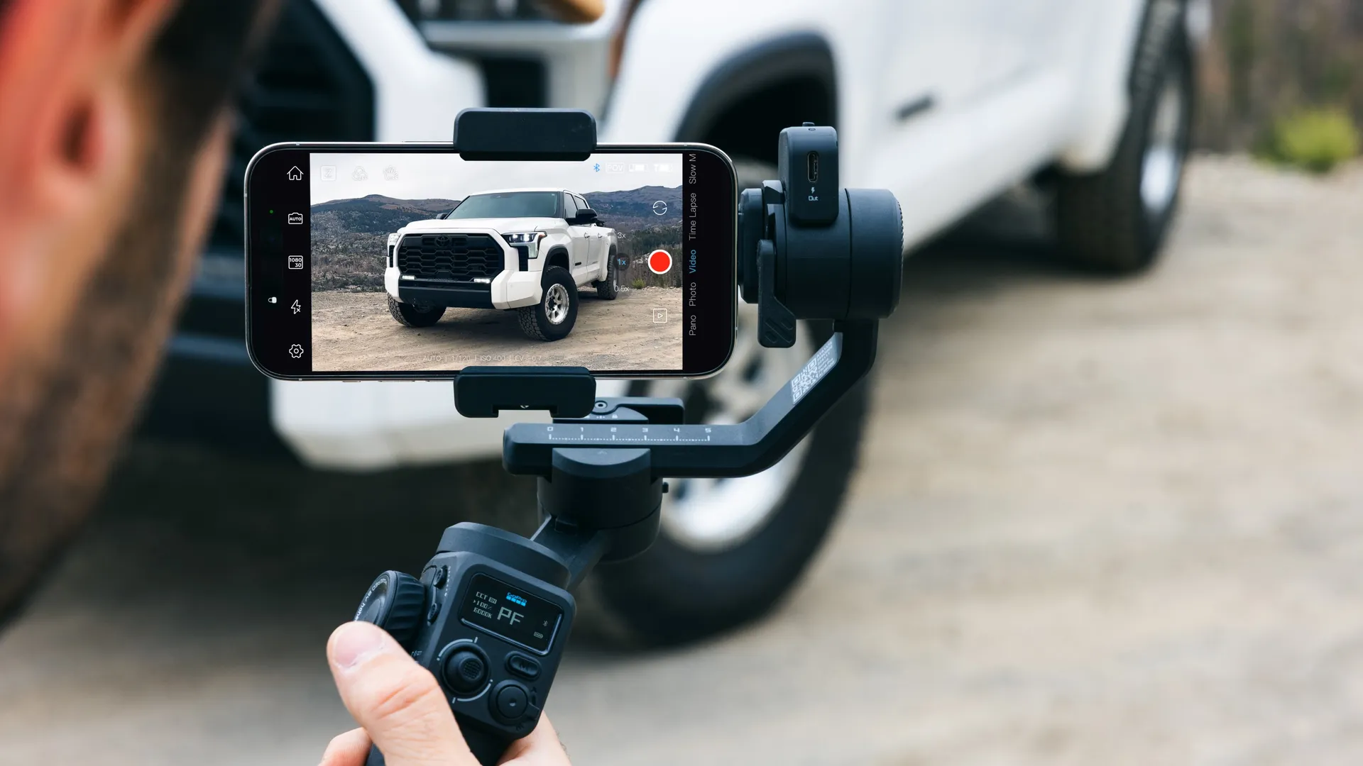 Eine Person filmt einen weißen Lkw mit einem auf einem Gimbal montierten Telefon. Der Lkw parkt auf einer unbefestigten Straße. Der Hintergrund ist unscharf.