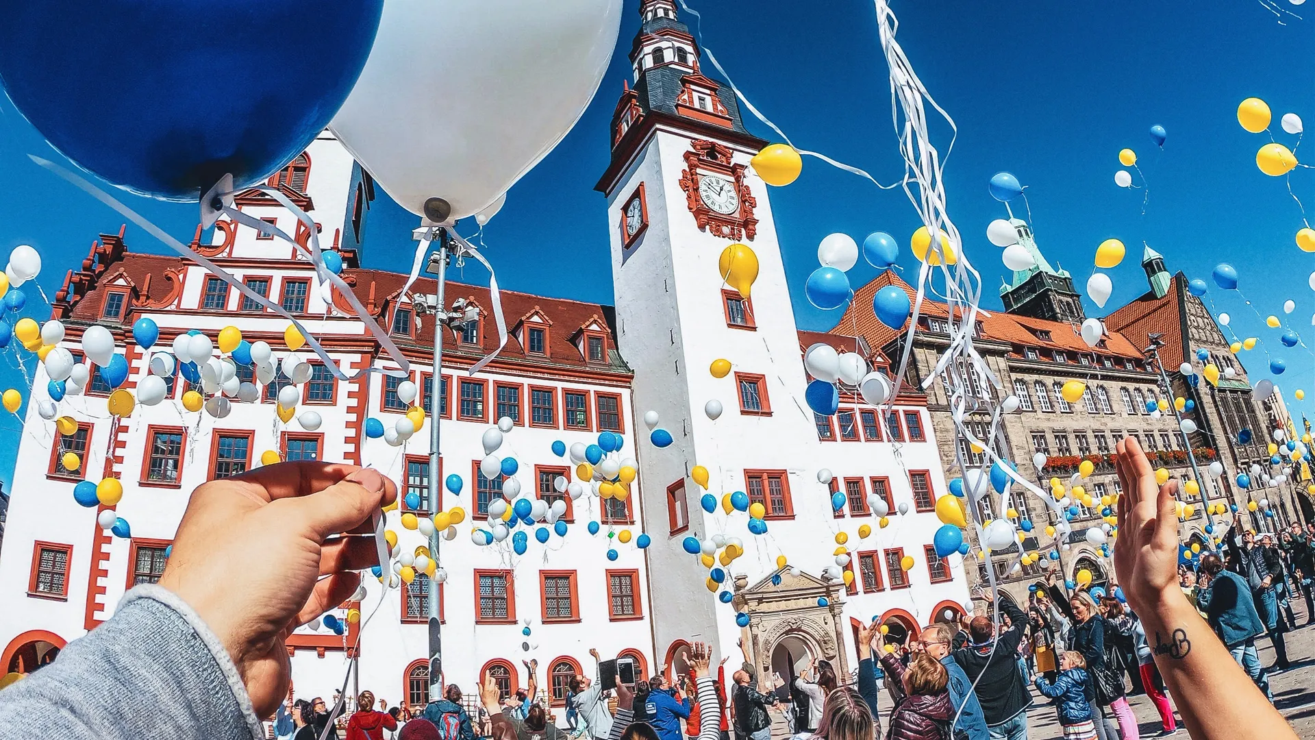 Persone rilasciano palloncini in una piazza. Un campanile ed edifici sullo sfondo. Cielo blu.