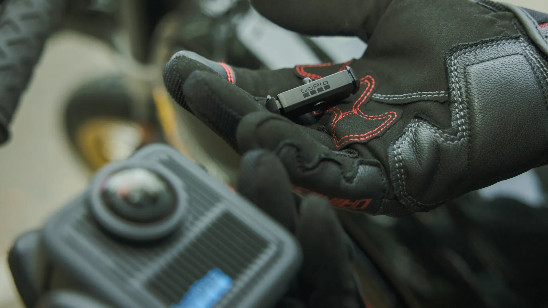 Ein schwarzer Handschuh hält eine GoPro-Halterung. Rote Nähte akzentuieren den Handschuh. Ein Motorradteil ist im verschwommenen Vordergrund.