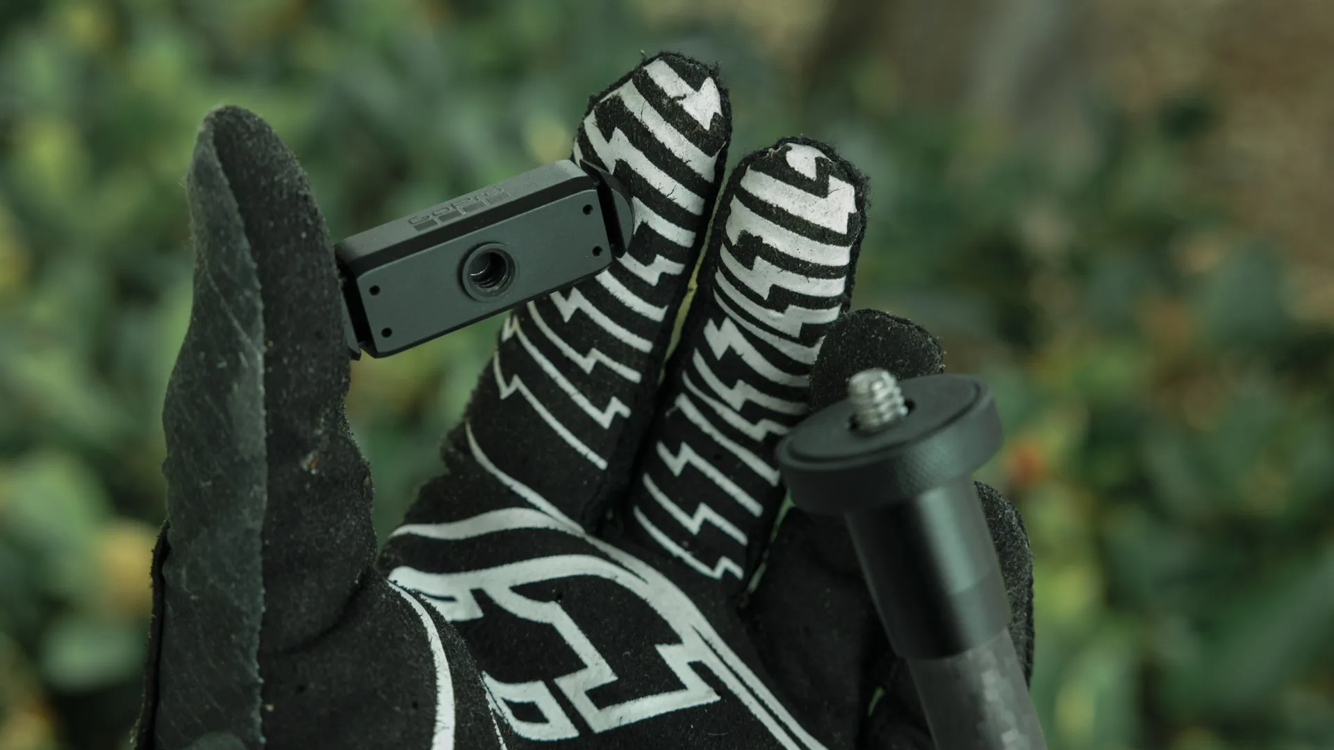 Hand mit schwarz-weißem Handschuh, hält eine schwarze Kamera und Stativhalterung. Grünes Laub im Hintergrund.
