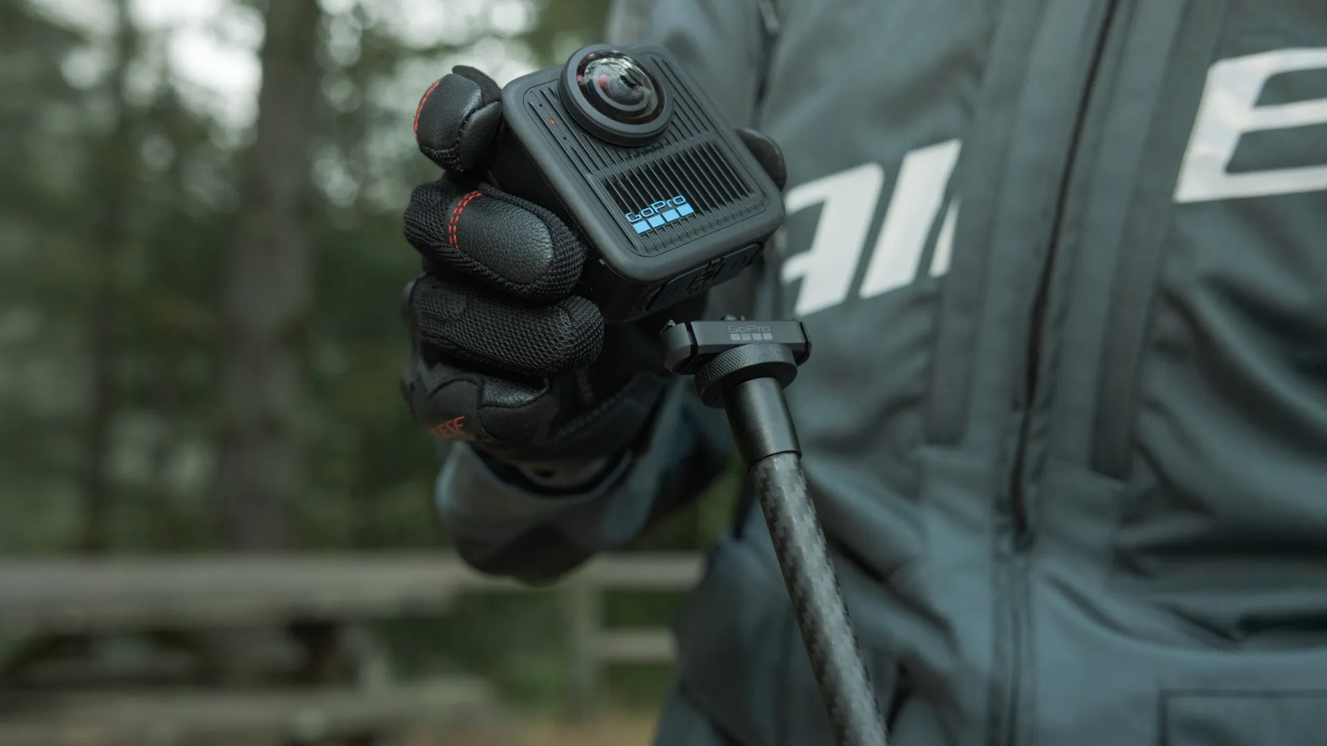 Eine Person hält eine schwarze GoPro-Kamera an einem Selfie-Stick. Sie trägt schwarze Handschuhe und eine graue Jacke. Der Hintergrund sind verschwommene Bäume.