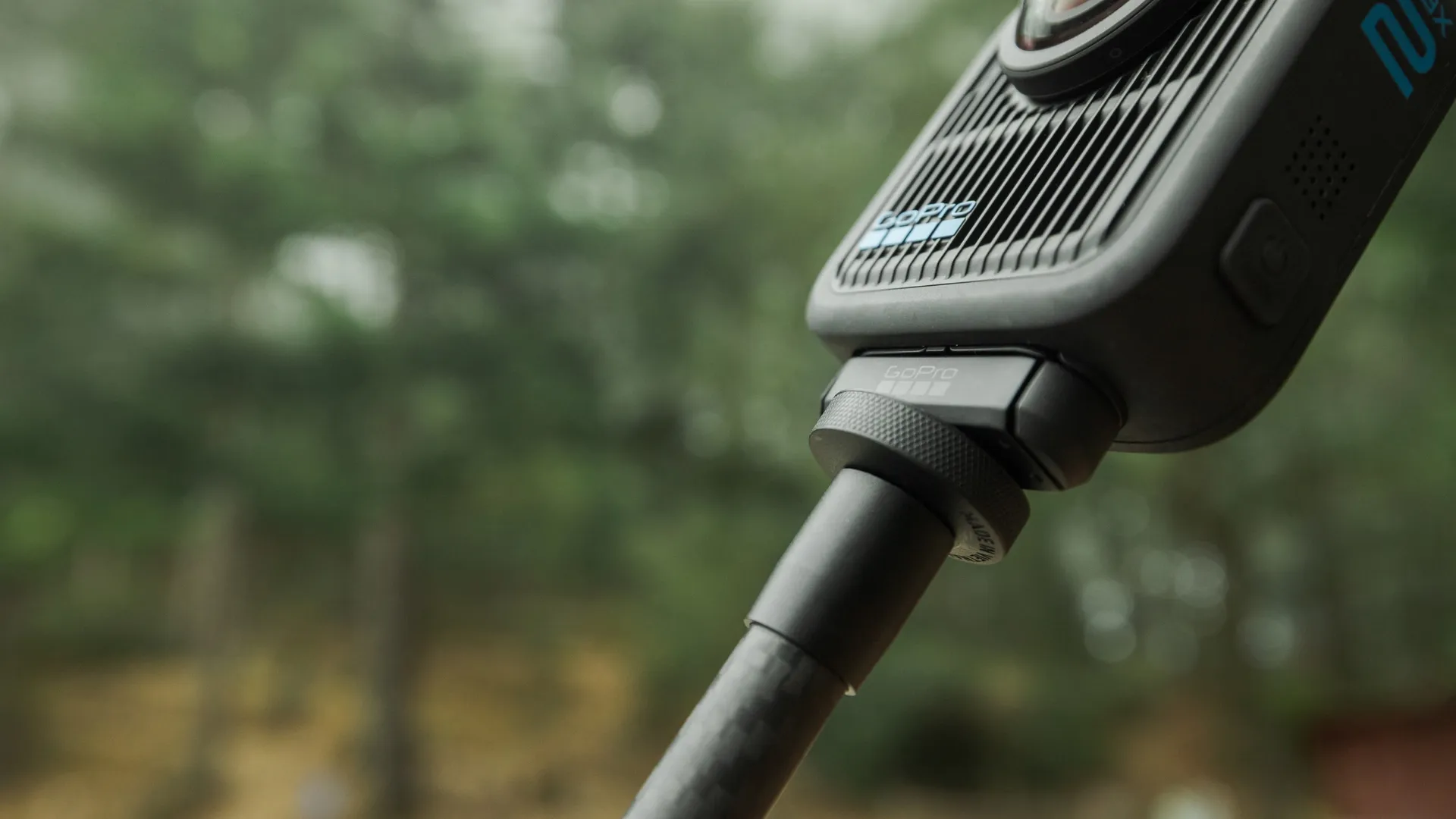 GoPro-Kamera an einem schwarzen Stab befestigt. Verschwommener grüner Hintergrund mit Bäumen. GoPro-Logo sichtbar.