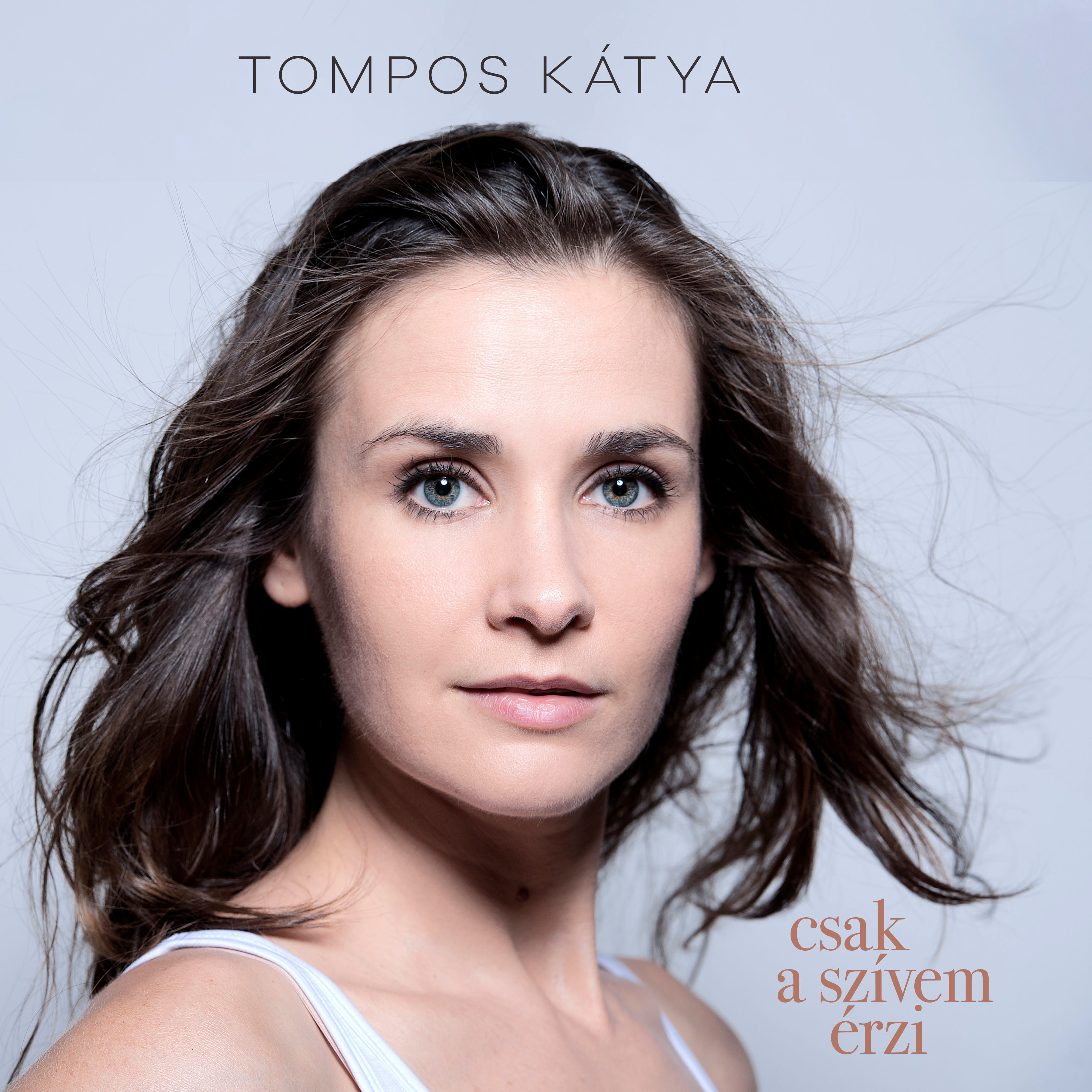 Egy nő a nézőre néz. Sötét haja hátrafelé van fújva. A szöveg a következőt írja: 'Tompos Katya'. A háttér világos.