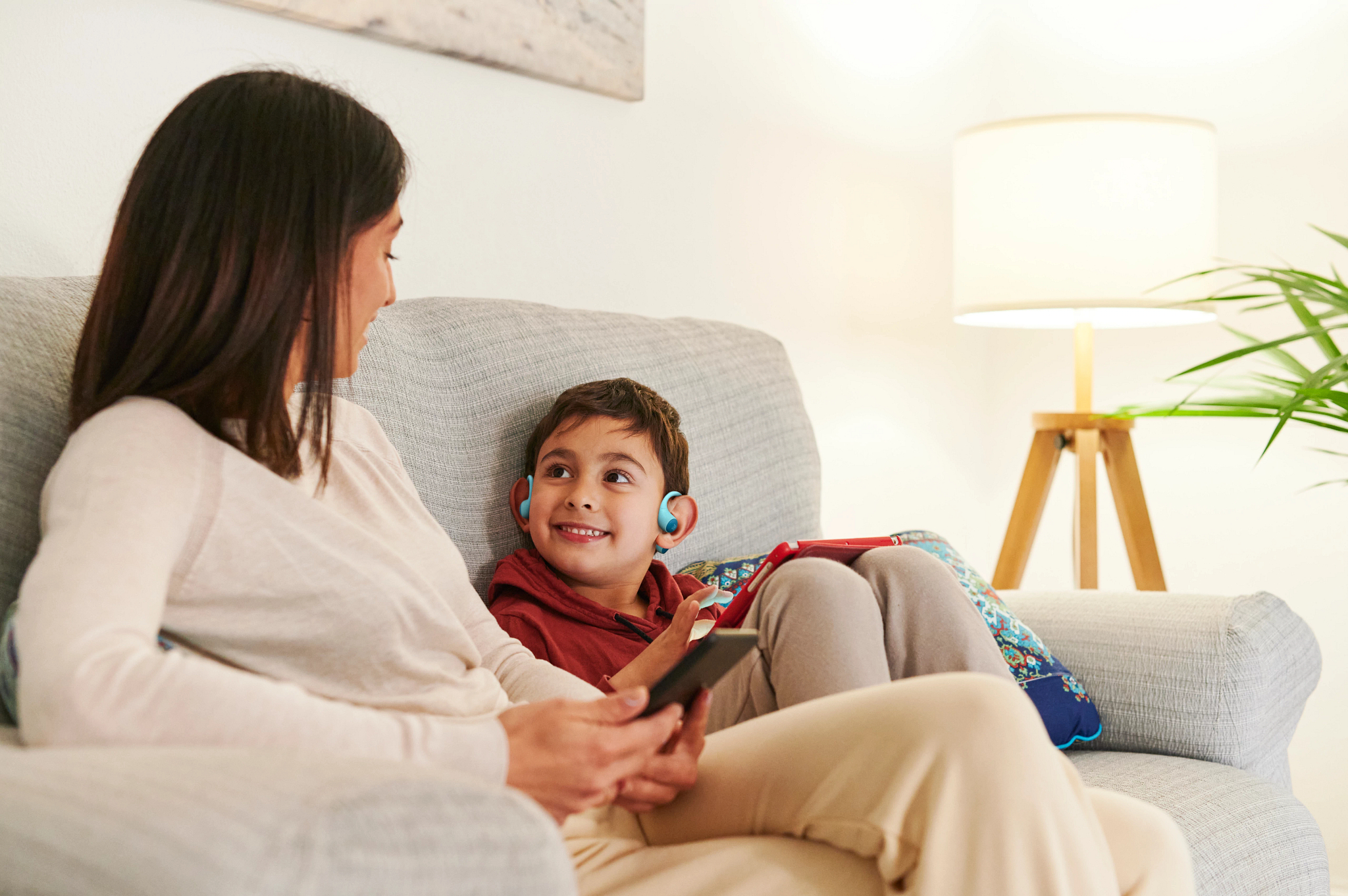 Eine Frau und ein Junge sitzen auf einem Sofa. Der Junge trägt blaue Hörgeräte. Eine Lampe und eine Pflanze sind im Hintergrund.
