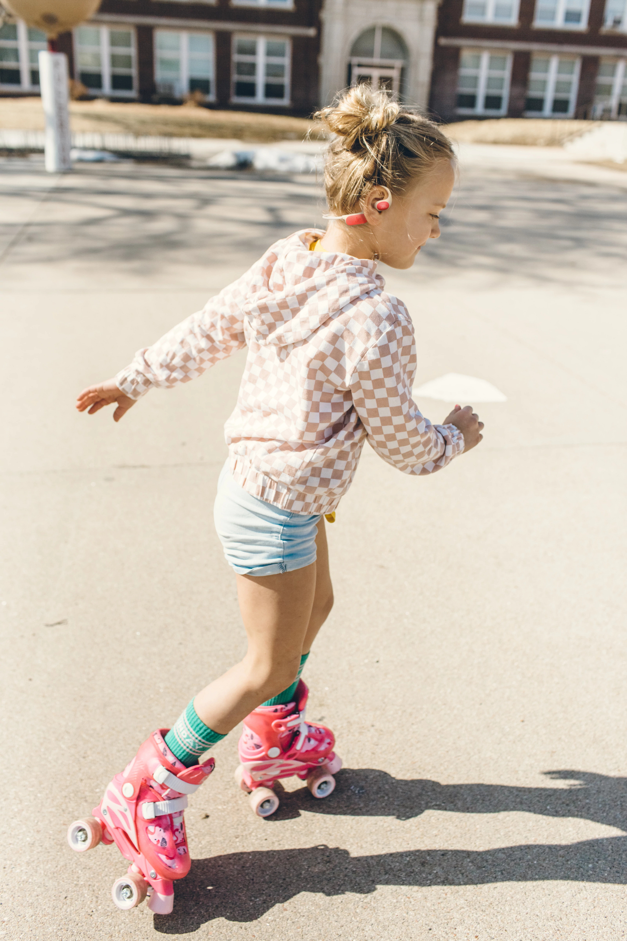 Ein Mädchen auf Rollschuhen trägt eine karierte Jacke und Shorts. Im Hintergrund ist ein Schulgebäude zu sehen.