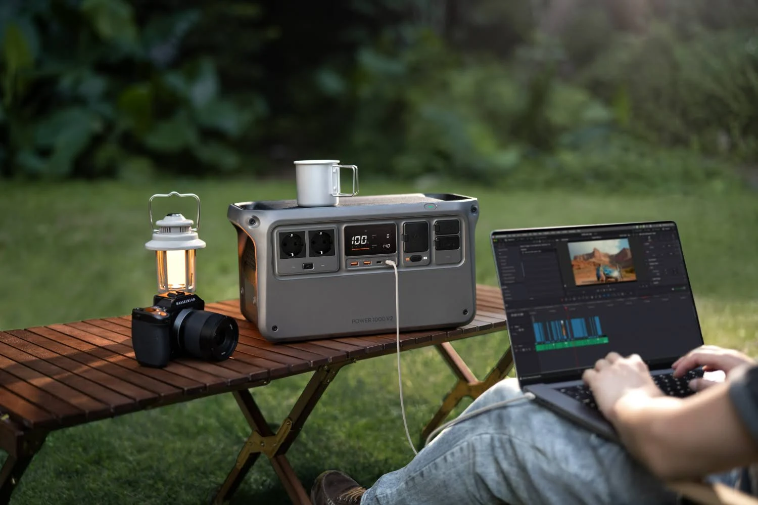 Una persona edita un video en una laptop al aire libre, con una estación de energía, cámara y linterna en una mesa.