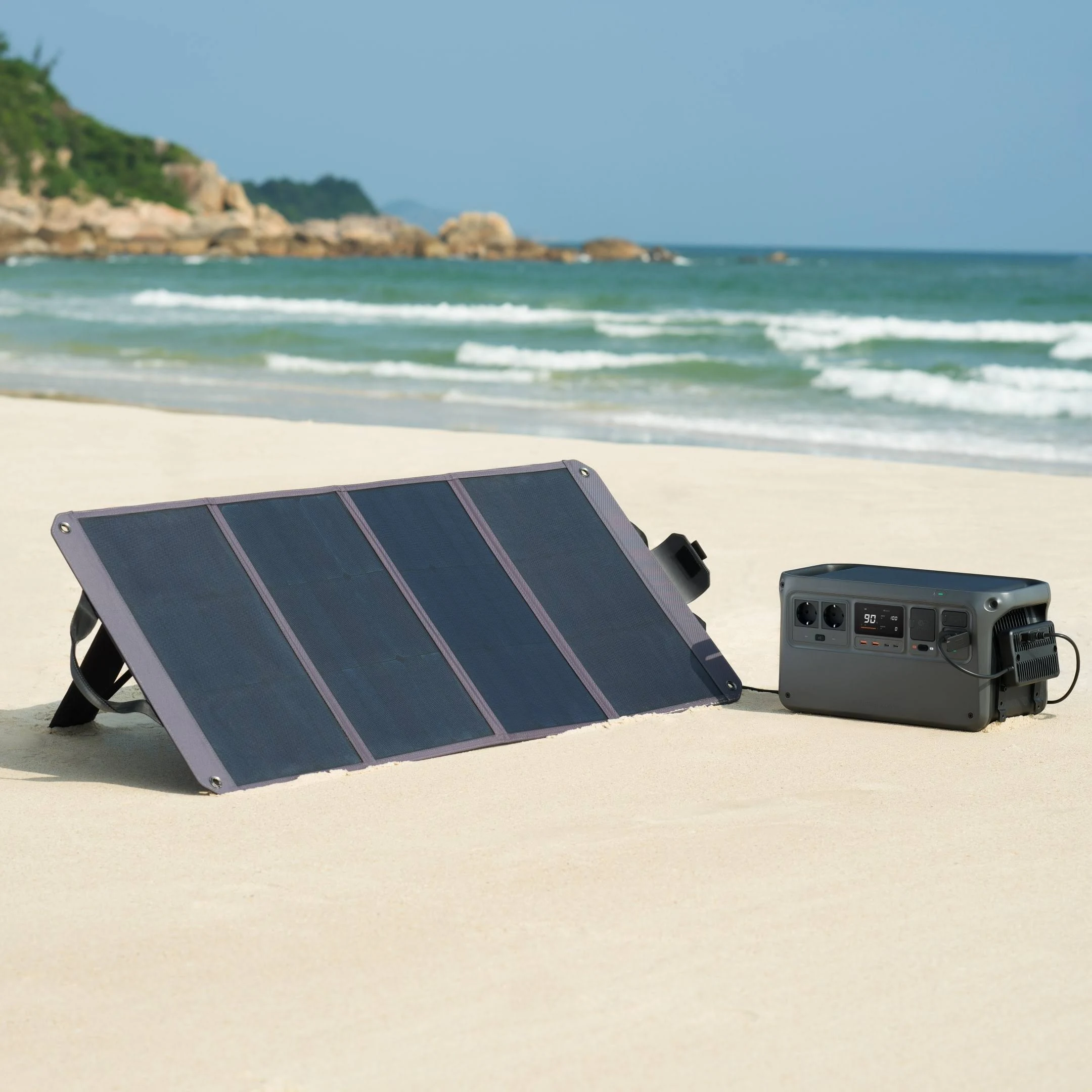 Un panel solar y una estación de energía están instalados en una playa arenosa, con el océano y rocas visibles.