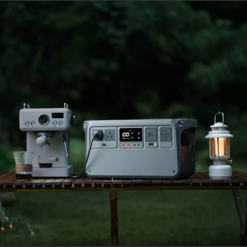 Una escena de camping con cafetera, estación de energía y linterna en una mesa de madera. Follaje verde en el fondo borroso.
