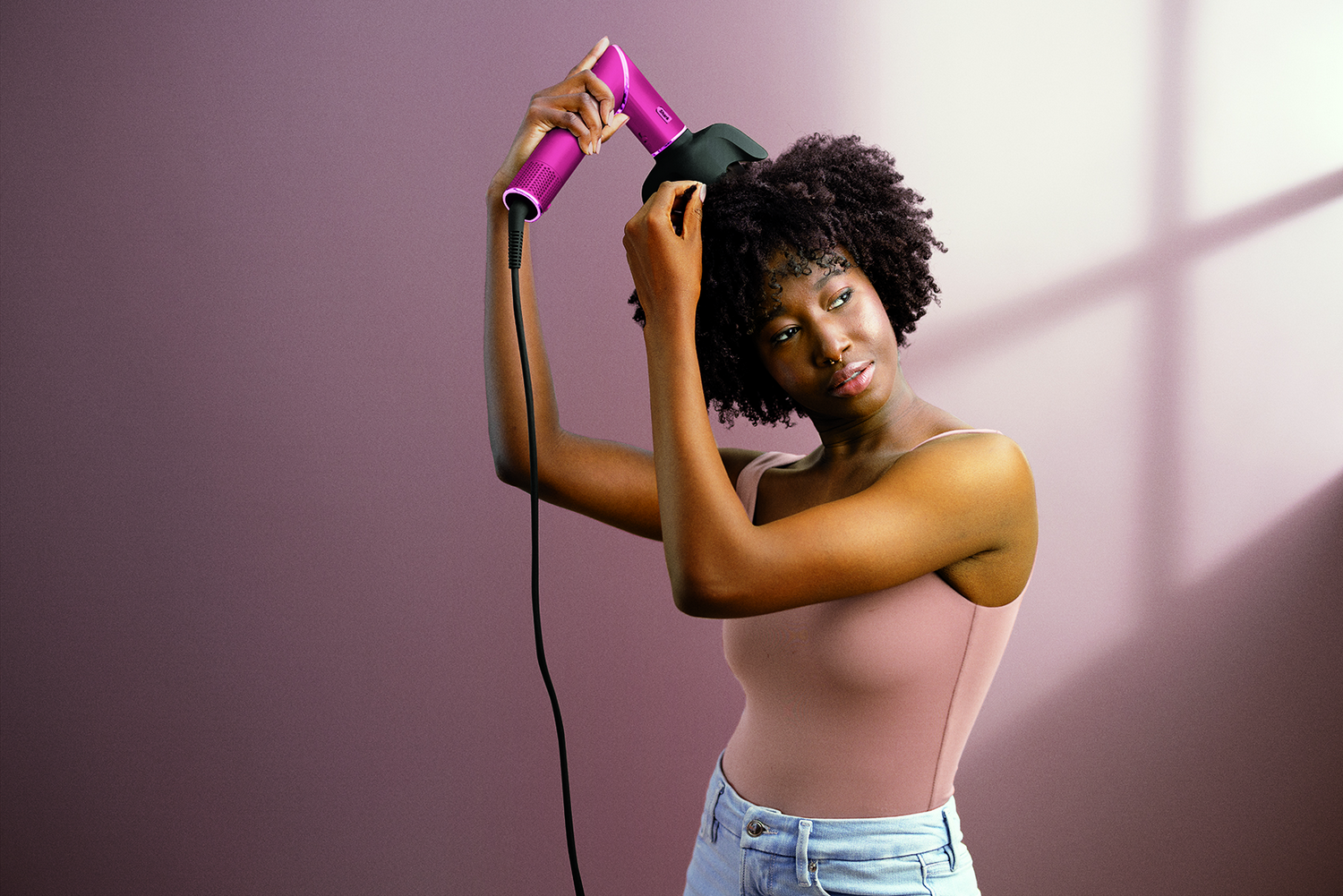 Una mujer con cabello rizado oscuro usa un secador de pelo rosa. Lleva un top rosa y vaqueros. El fondo es morado.