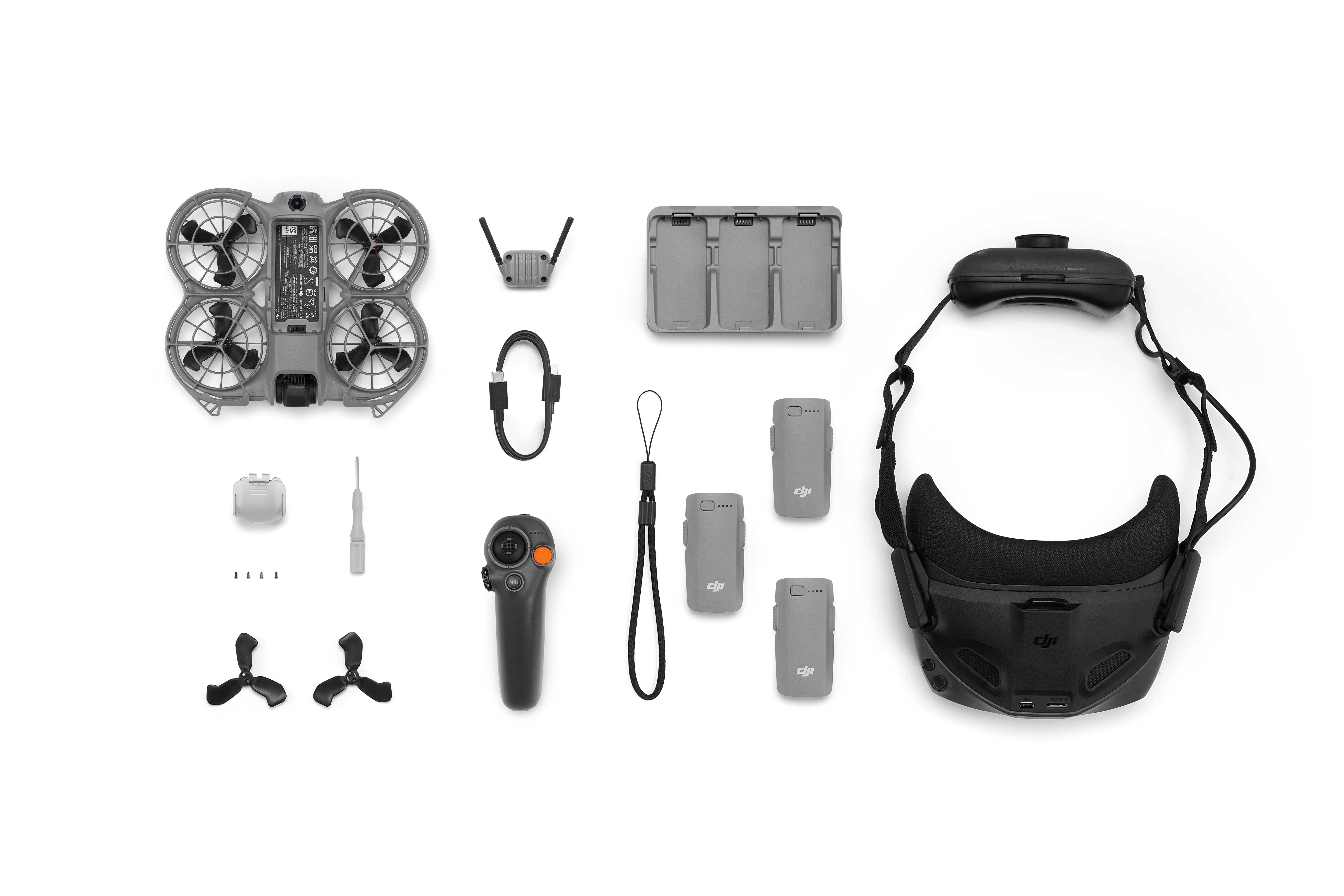 Drohnenteile auf weißer Oberfläche: Drohne, Brille, Batterien, Ladegerät, Fernbedienung, Kabel, Antennen und Propeller.