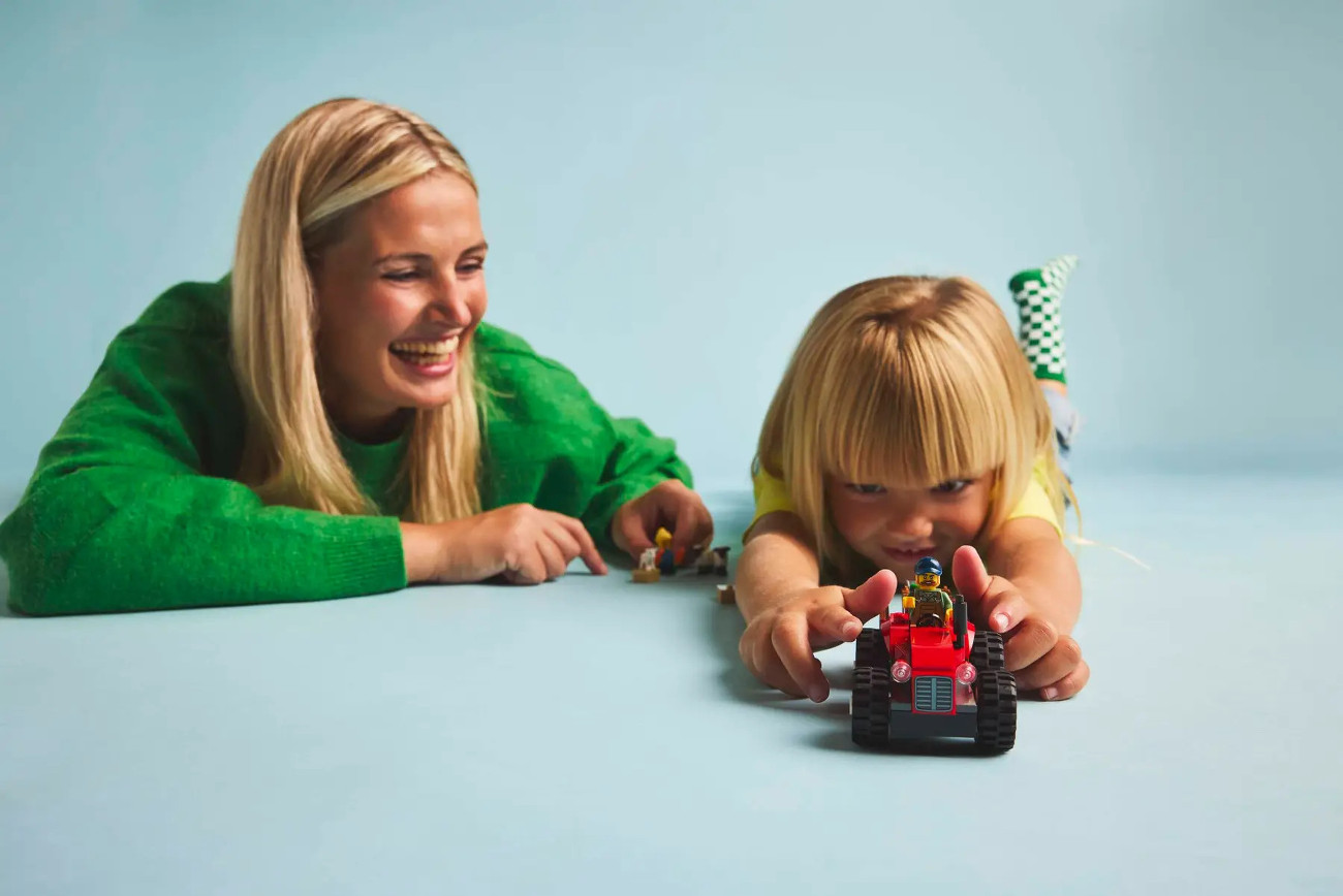 Kobieta i dziecko bawią się klockami Lego. Dziecko trzyma czerwony traktor Lego. Kobieta się uśmiecha.