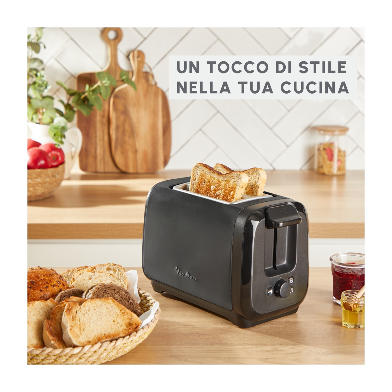 Un tostapane nero con pane tostato all'interno. Un cestino di fette di pane e vasetti di marmellata sono sulla superficie di legno.