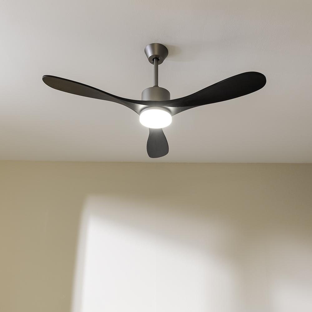 Un ventilador de techo moderno con tres aspas negras, una luz y un acabado metálico, contra un techo blanco y una pared beige.