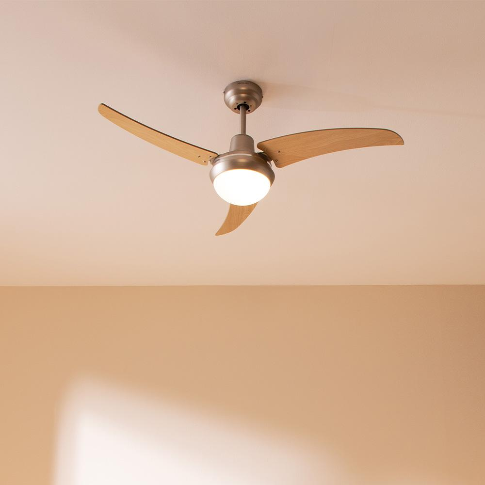 Un ventilador de techo con tres aspas de madera y una lámpara central. El ventilador está montado en un techo blanco.
