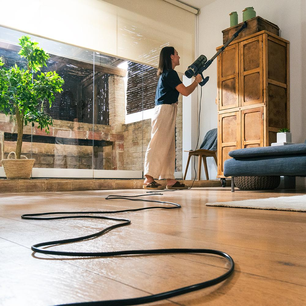 Mujer aspira una habitación. Lleva una camisa azul marino y pantalones beige. Hay un árbol, un armario y un sofá en la habitación.