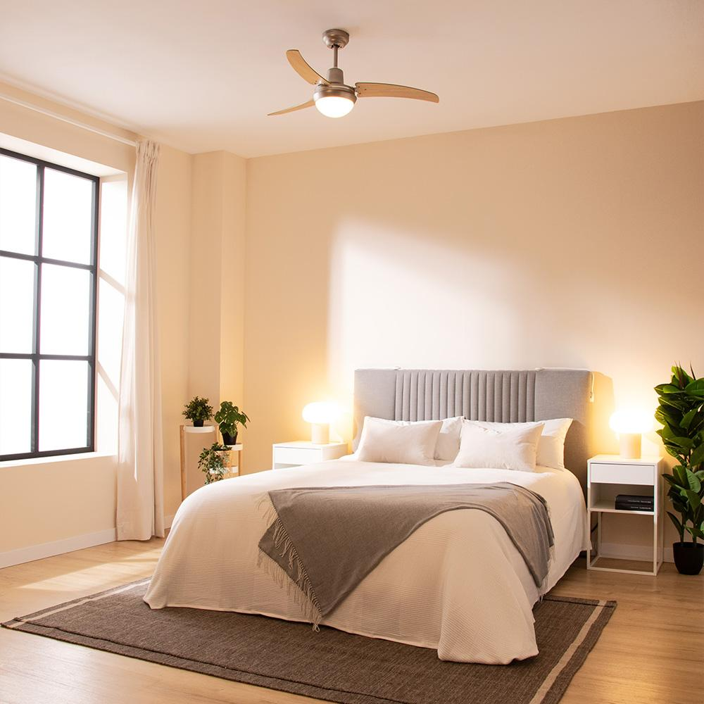 Un dormitorio con cama, dos mesitas de noche, una ventana grande y un ventilador de techo. La cama tiene un cabecero gris y una manta marrón.