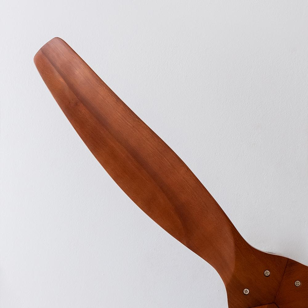 Una hélice de avión de madera está en ángulo sobre un fondo blanco. La hélice es marrón con detalles de tornillos en la base.