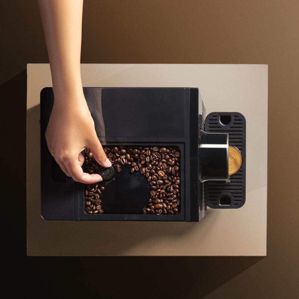 Una mano coloca una tapa negra en una cafetera llena de granos de café. Se ve una cápsula de café.