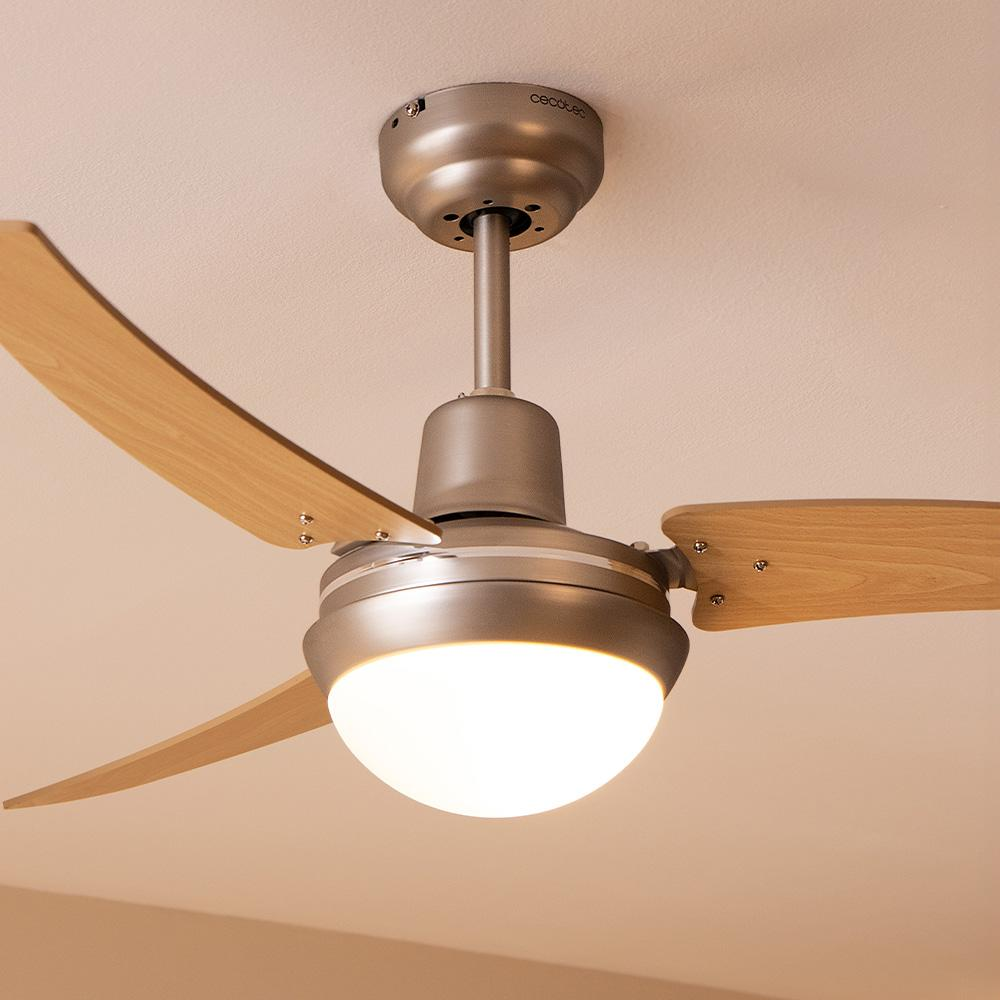 Un ventilador de techo con aspas de madera y una luz. El ventilador es de color plateado y está montado en un techo blanco. La luz está encendida.