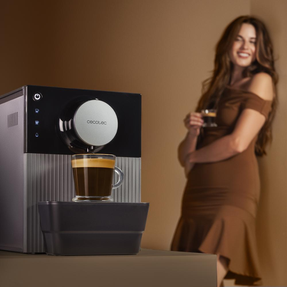 Cafetera con vaso de café delante. Una mujer con vestido marrón, a un lado, sostiene un vaso pequeño.