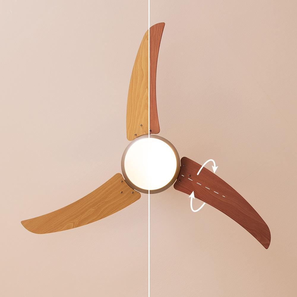 Un ventilador de techo con tres aspas de madera. Las aspas son de diferentes tonos marrones. Una luz está en el centro. Flechas muestran movimiento.