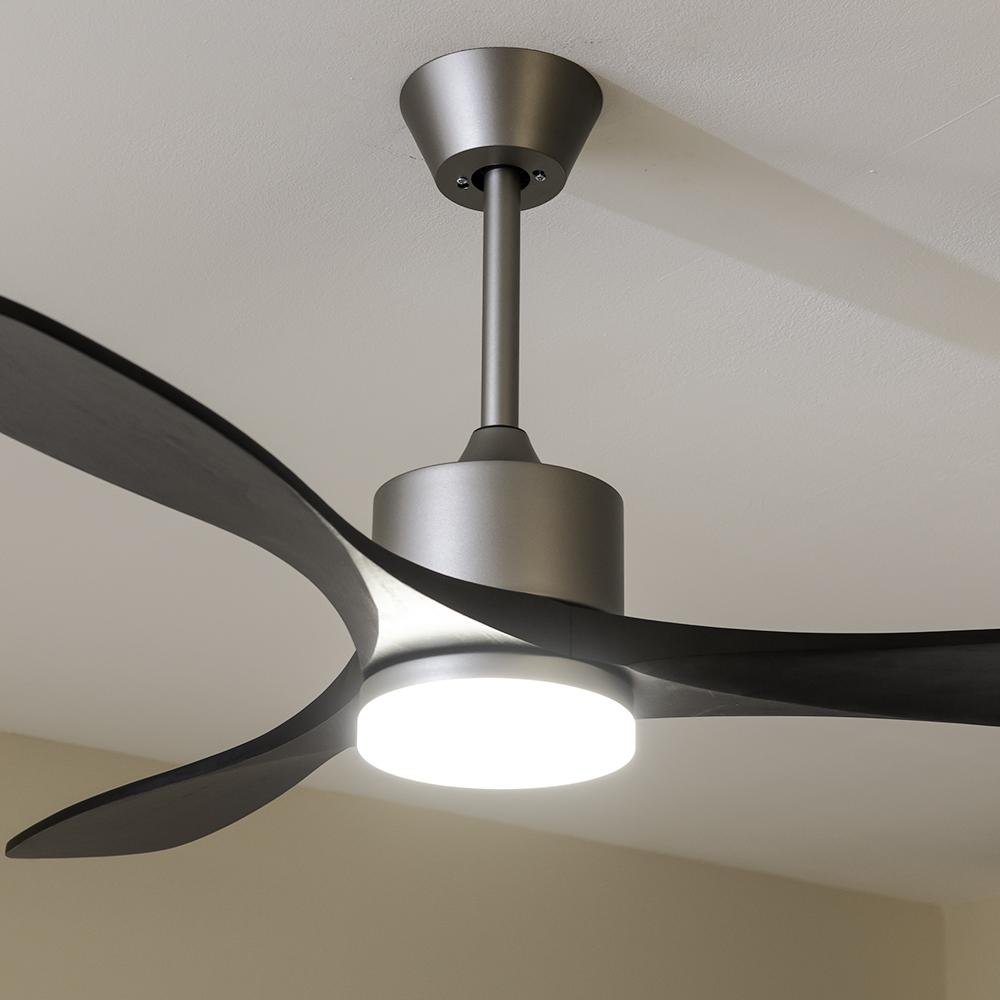 Un ventilador de techo con tres aspas oscuras y una lámpara central está montado en un techo blanco. El ventilador tiene un acabado metálico.