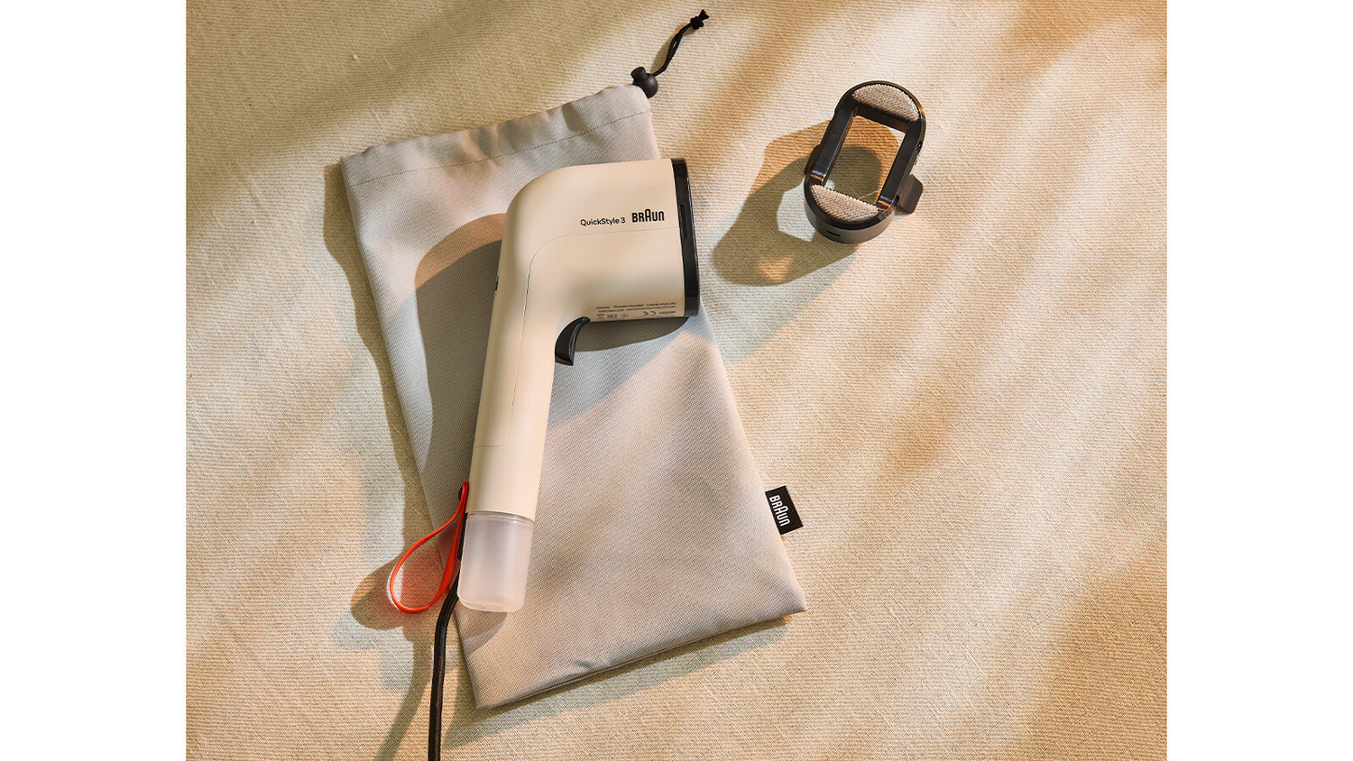 Una plancha de ropa Braun QuickStyle 3 crema, dos bolsas grises y un cabezal de limpieza negro sobre tela beige.