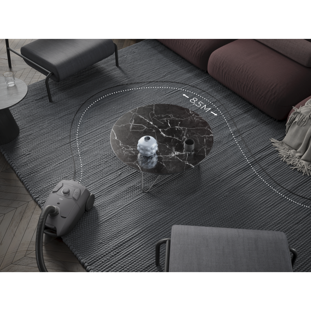 A gray vacuum cleaner on a gray rug. A marble table, sofa, and chairs are in the background. A dotted line shows the vacuum's reach.