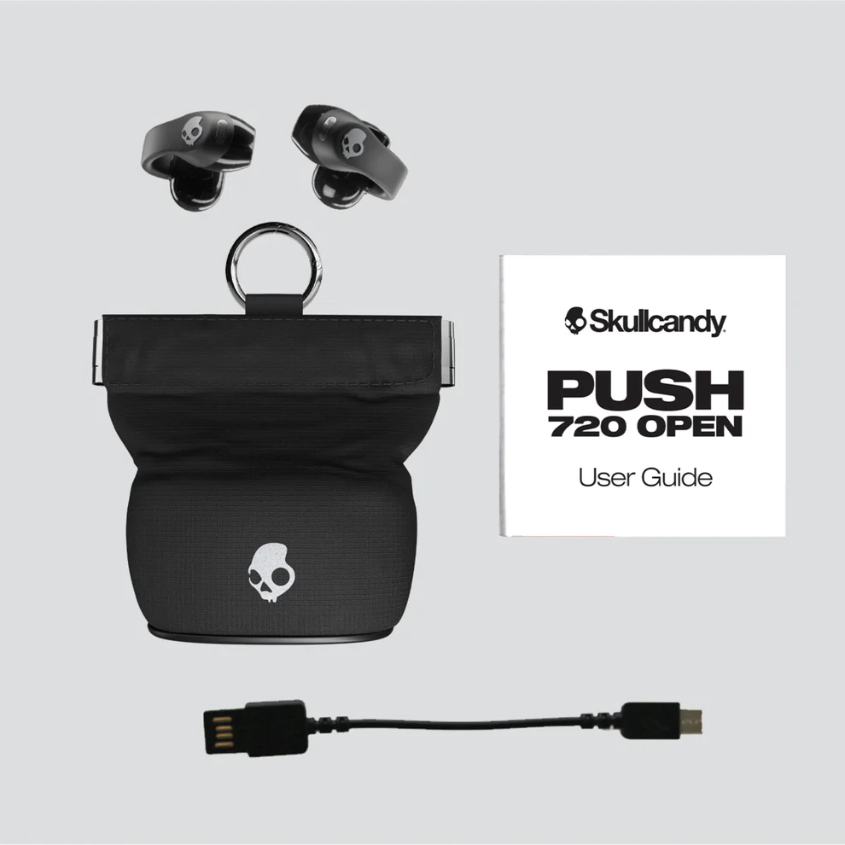 Schwarze Skullcandy-Ohrhörer, Ladeetui, Benutzerhandbuch und ein USB-Kabel auf hellgrauem Hintergrund. Das Etui hat ein Totenkopf-Logo.