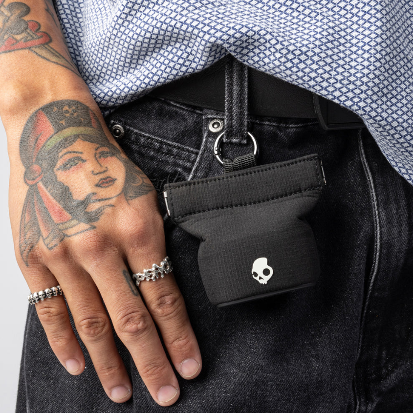 Eine Person mit Tätowierungen und Ringen hält eine schwarze Tasche mit einem Totenkopf-Logo an ihrer Jeans. Ein blaues Hemd ist sichtbar.
