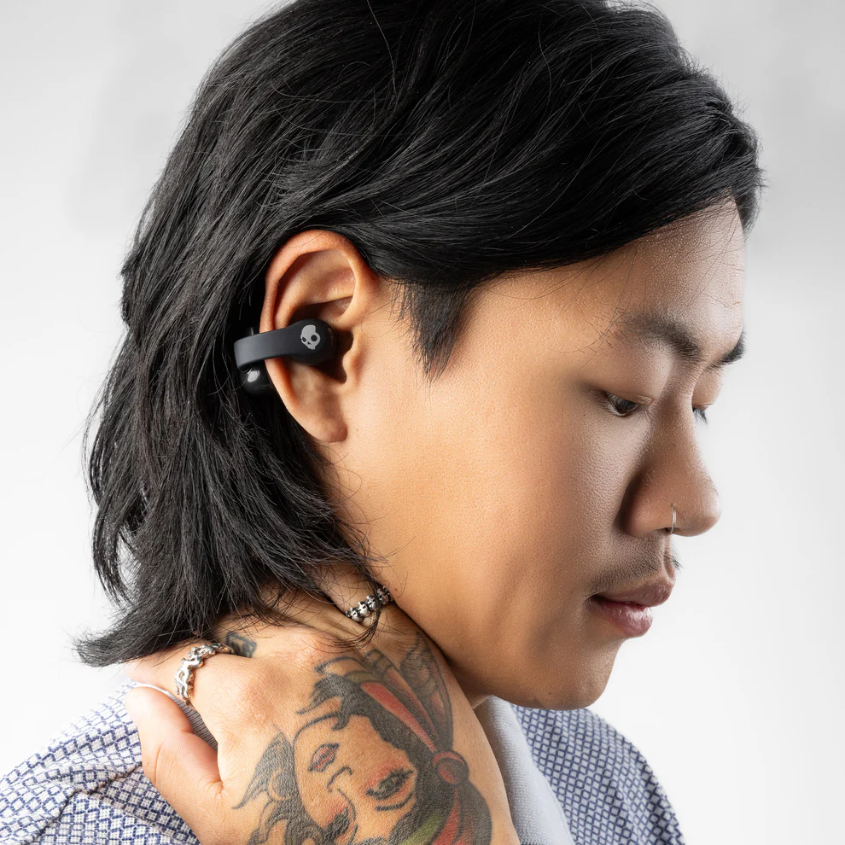 Ein Mann mit langen schwarzen Haaren trägt einen schwarzen Ohrhörer. Er hat ein Tattoo auf dem Arm und einen Nasenring.