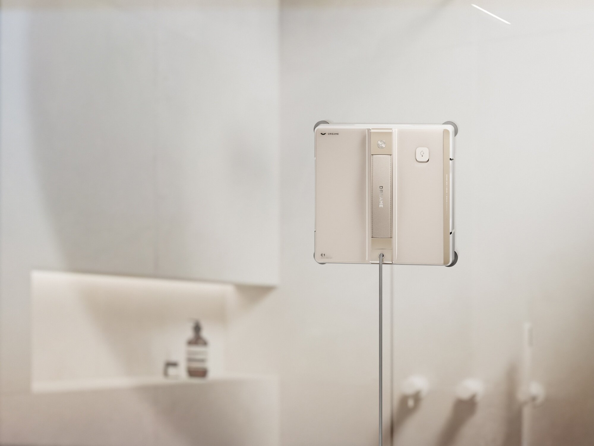 Beżowy robot do czyszczenia okien przymocowany do szklanej powierzchni. W tle półka z butelką i mydłem.