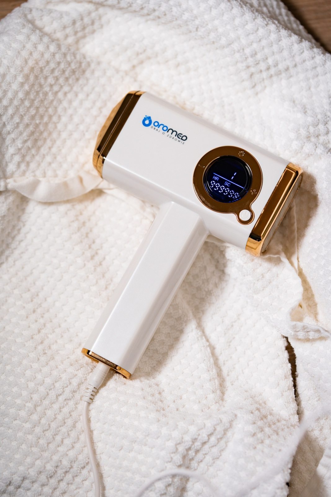 Biało-złote urządzenie do usuwania włosów na białym ręczniku. Urządzenie ma wyświetlacz i jest podłączone.