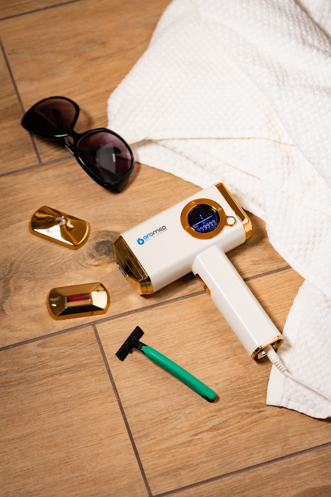 Białe i złote urządzenie do usuwania włosów, okulary przeciwsłoneczne, złote płytki, zielona maszynka do golenia i biały ręcznik na drewnianej powierzchni.