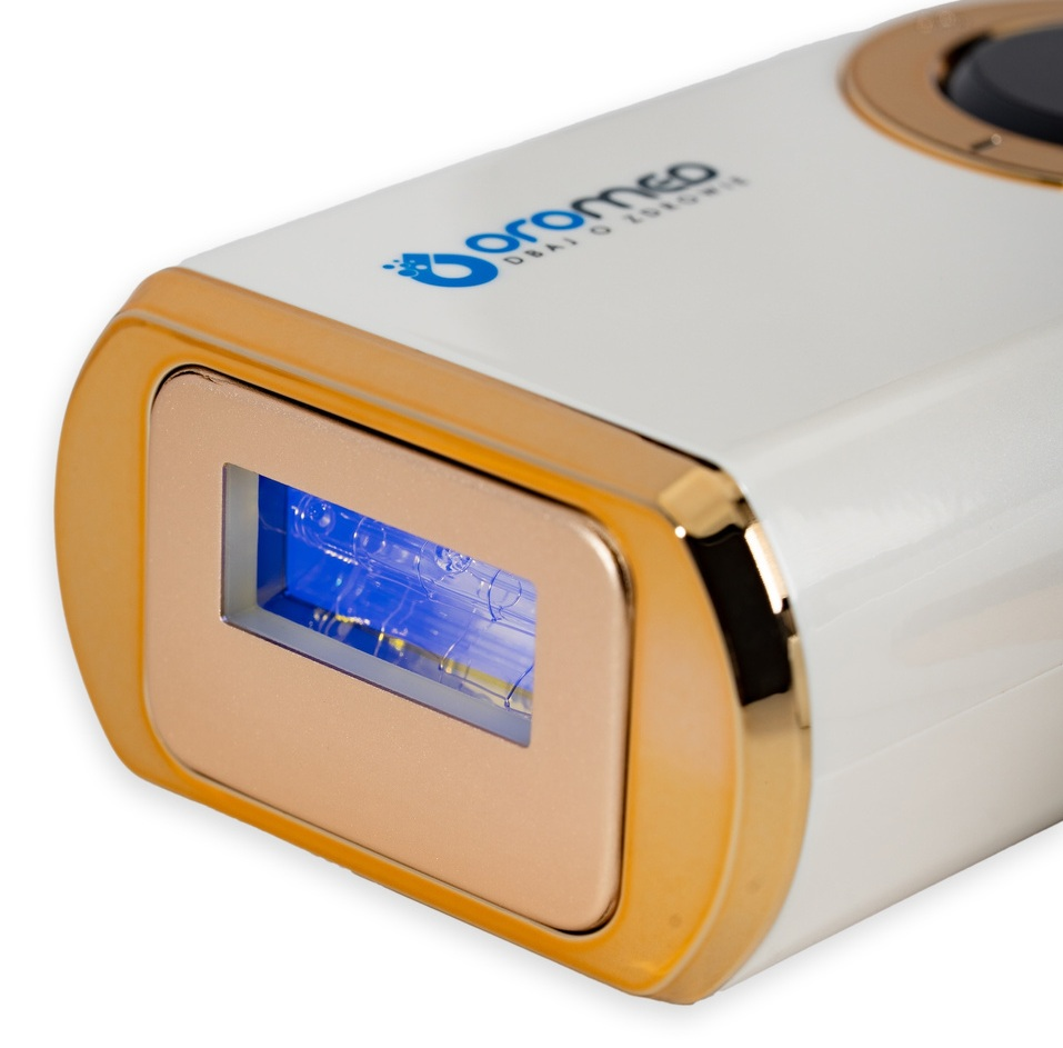 Białe i złote urządzenie do usuwania włosów laserem. Ma złotą ramkę wokół obszaru emitującego niebieskie światło. Marka jest widoczna.