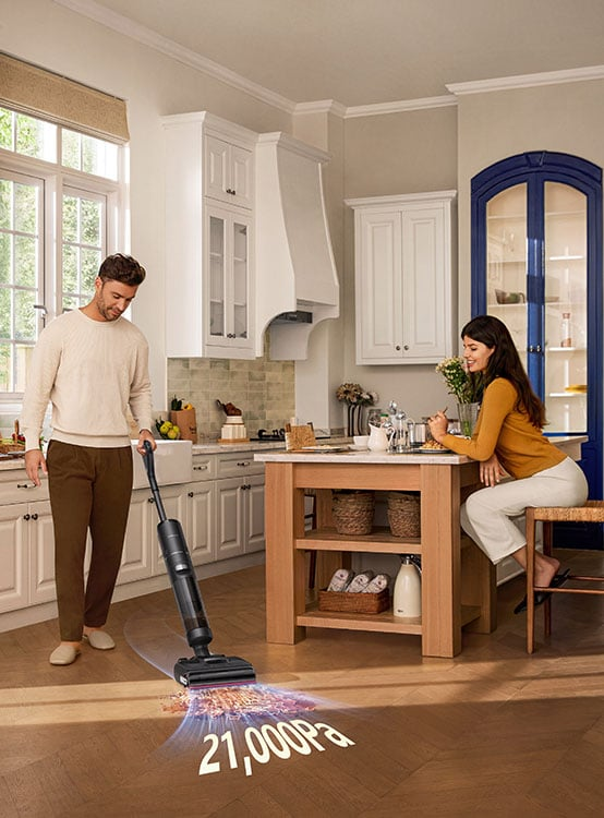 Mężczyzna odkurza podłogę, a kobieta je przy wyspie kuchennej. Odkurzacz pokazuje 21 000 Pa. Kuchnia z białymi szafkami.