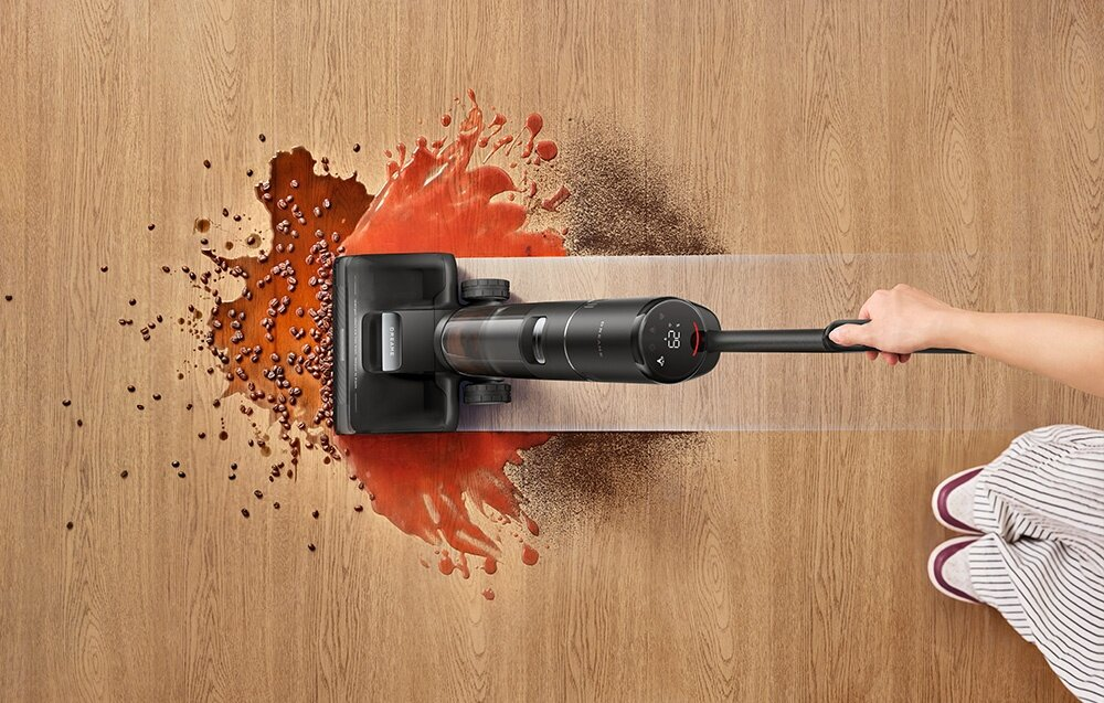 Czarny odkurzacz czyści rozlaną kawę na drewnianej podłodze. Ręka trzyma odkurzacz. Widoczne są stopy osoby w paski.