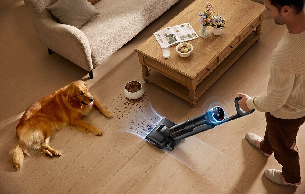 Mężczyzna odkurza podłogę, golden retriever leży. W pobliżu miska z rozsypaną karmą dla psów. Widoczne są sofa i stół.